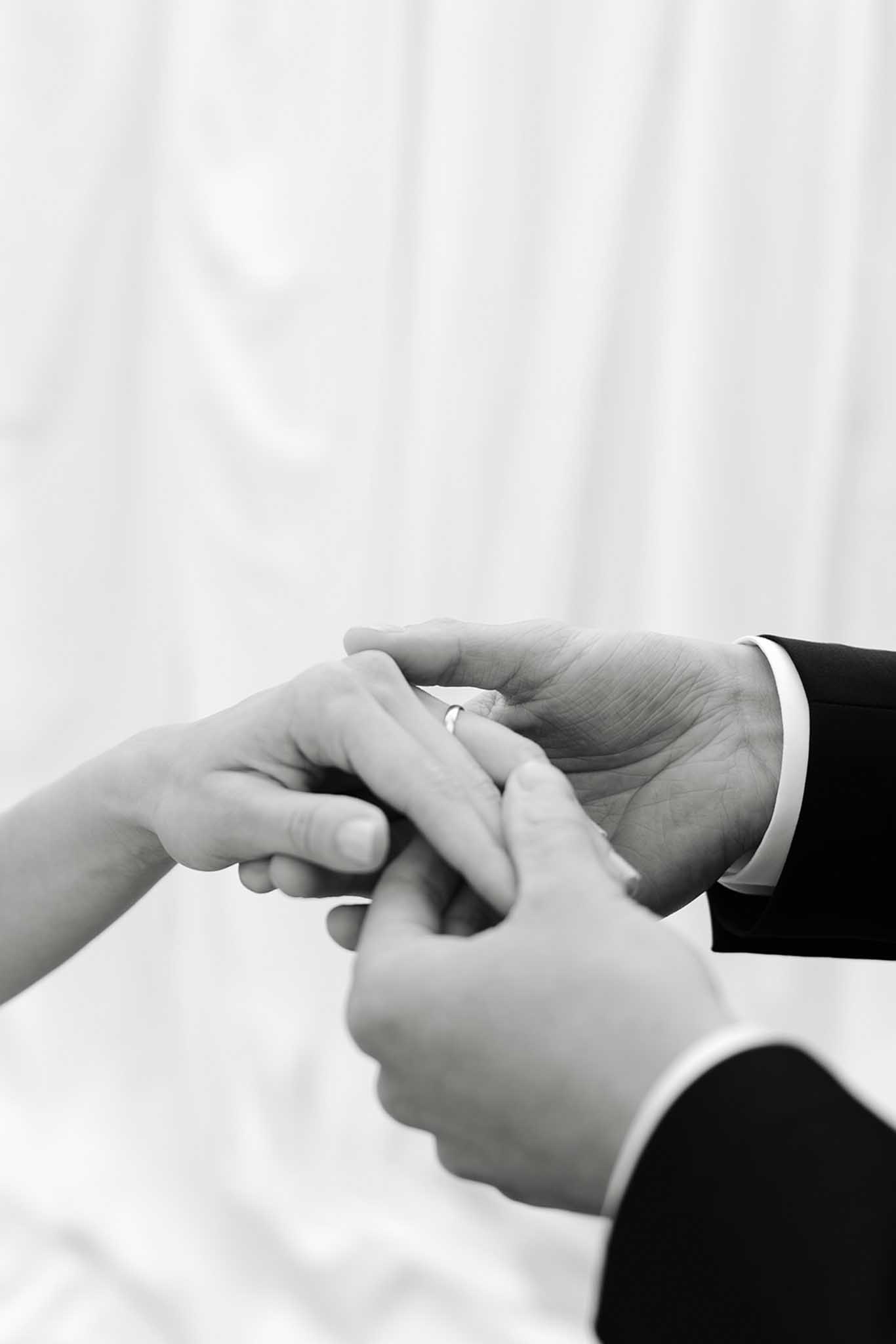 Close-up of bride and groom's hands during ring exchange, black and white, dark suit cuff against white dress
