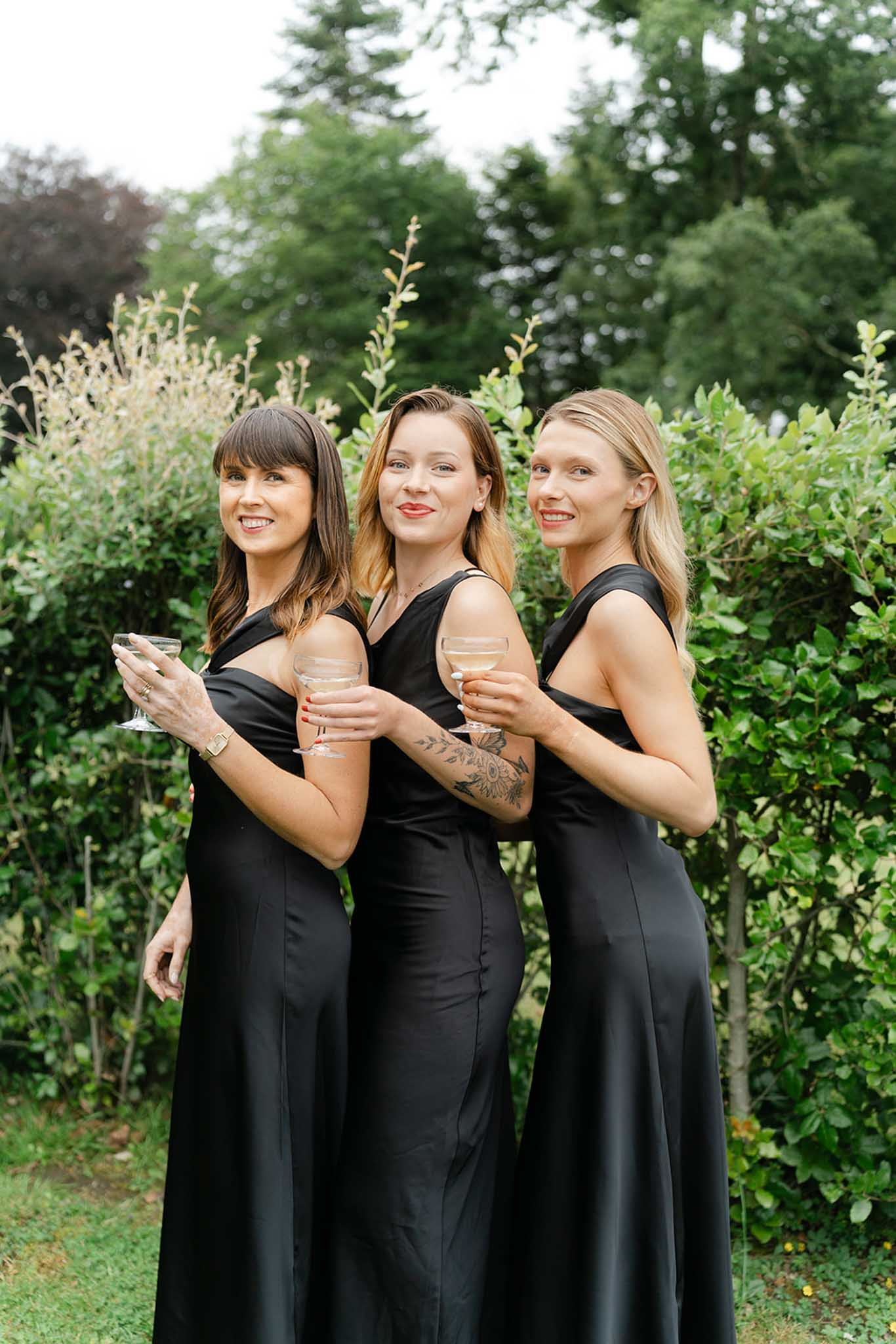 Three bridesmaids in matching black floor-length dresses holding champagne in garden