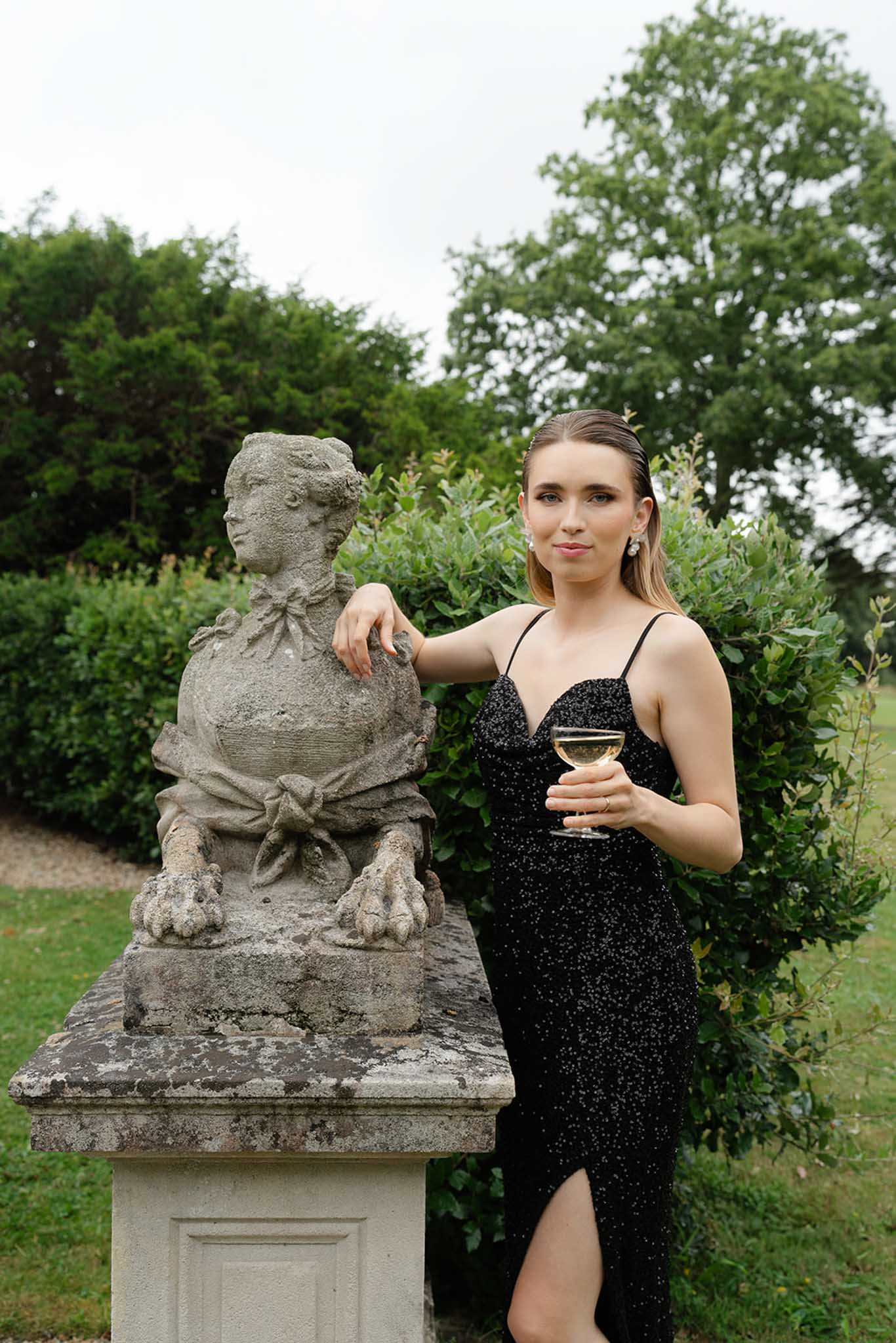 Guest in black beaded evening gown holding champagne coupe beside stone bust sculpture in formal garden