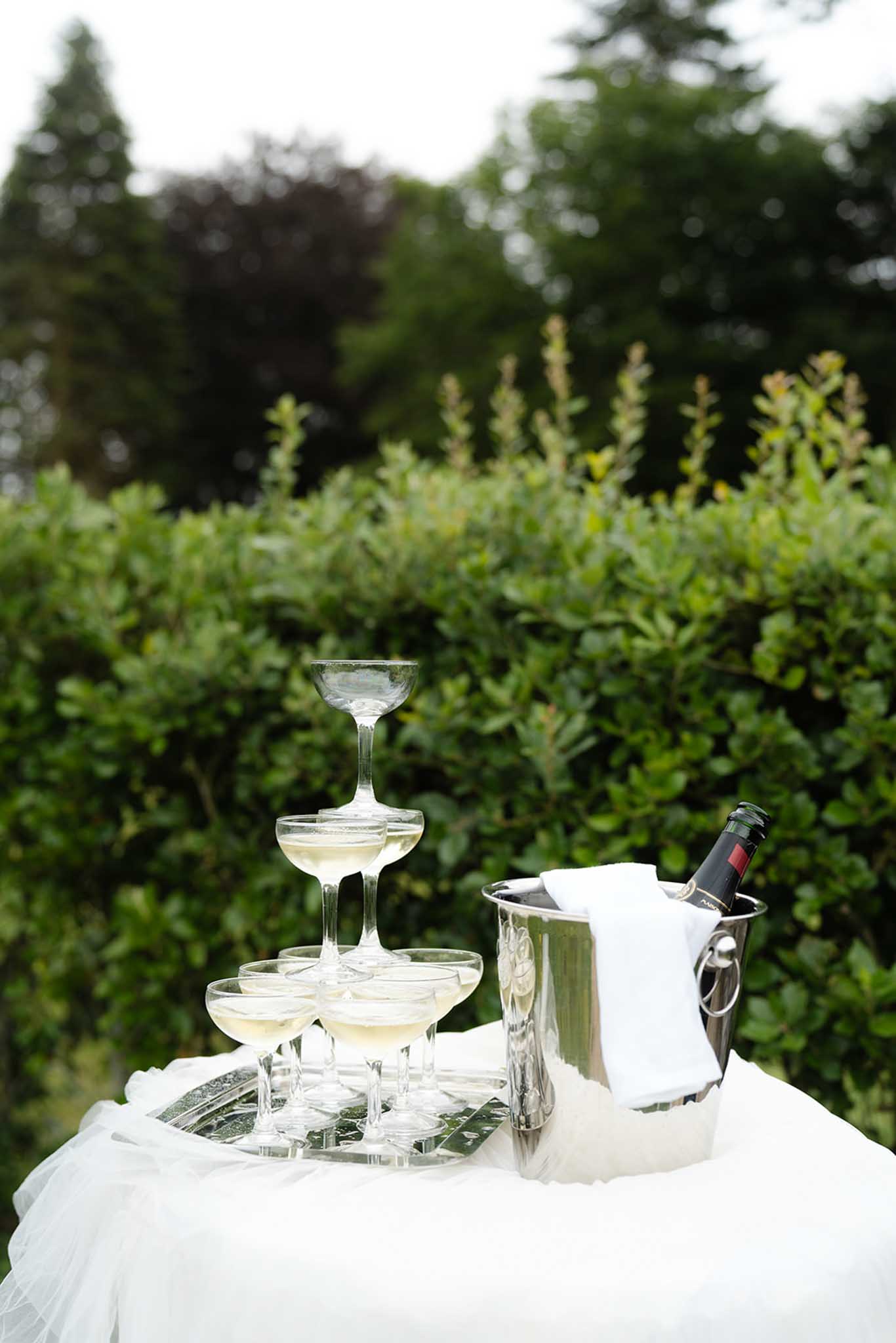 Champagne tower of coupe glasses on white-draped table in a formal garden with clipped hedges