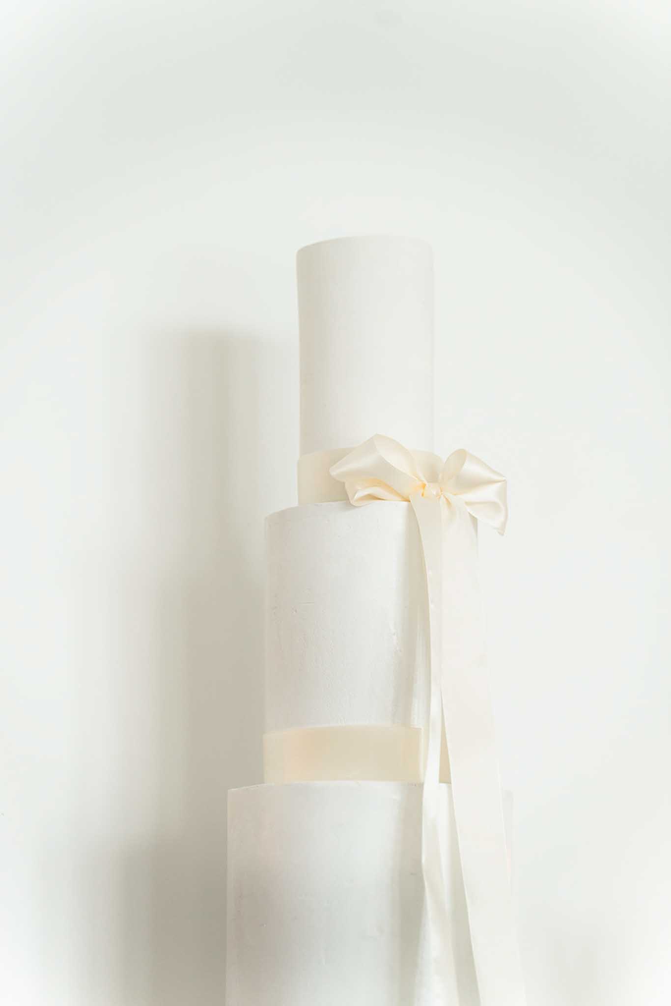 Four-tier white wedding cake decorated with single cream silk ribbon bow, minimal styling, even soft lighting