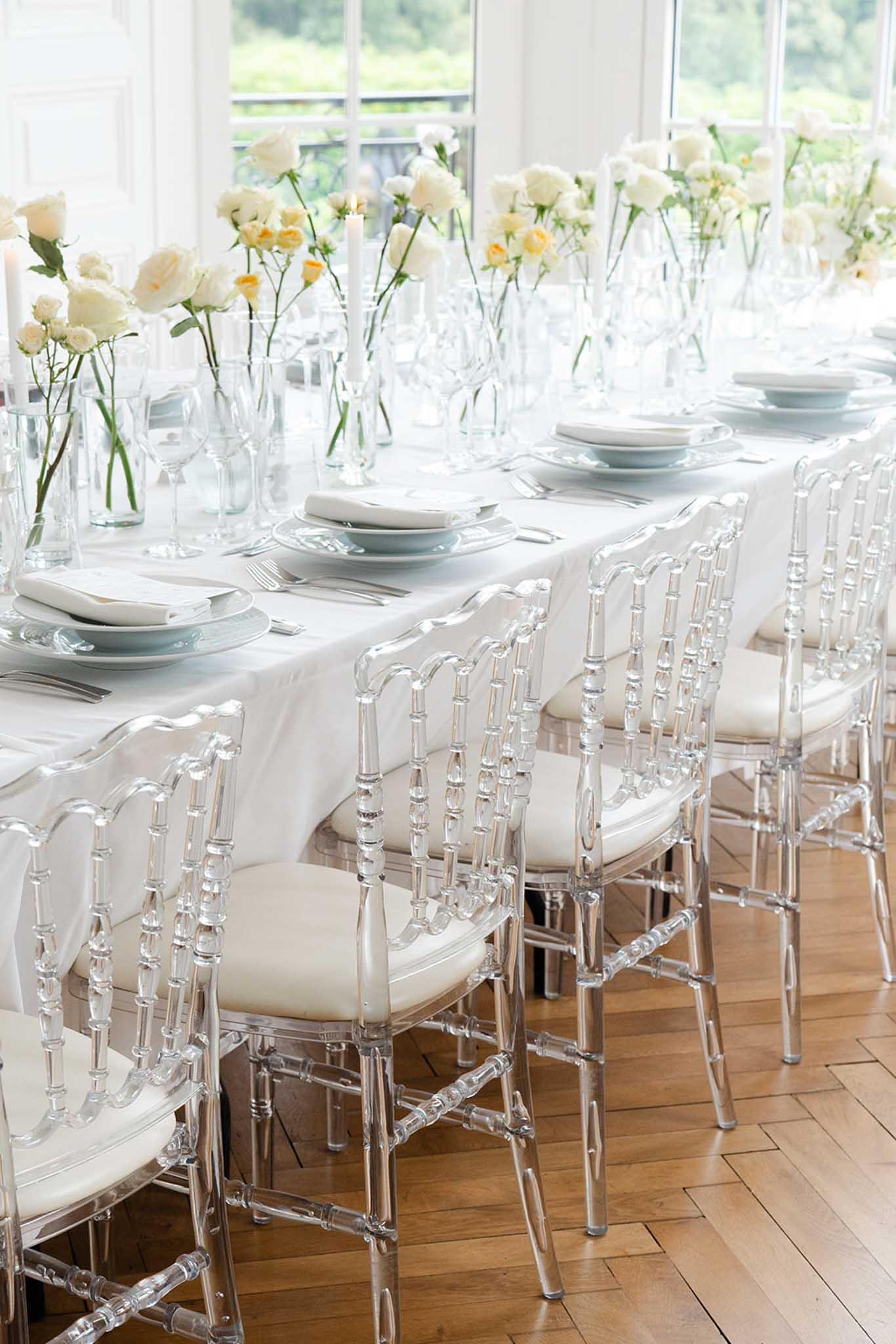 Long reception table with pale blue and white plates, clear acrylic chairs and ivory carnation centerpieces