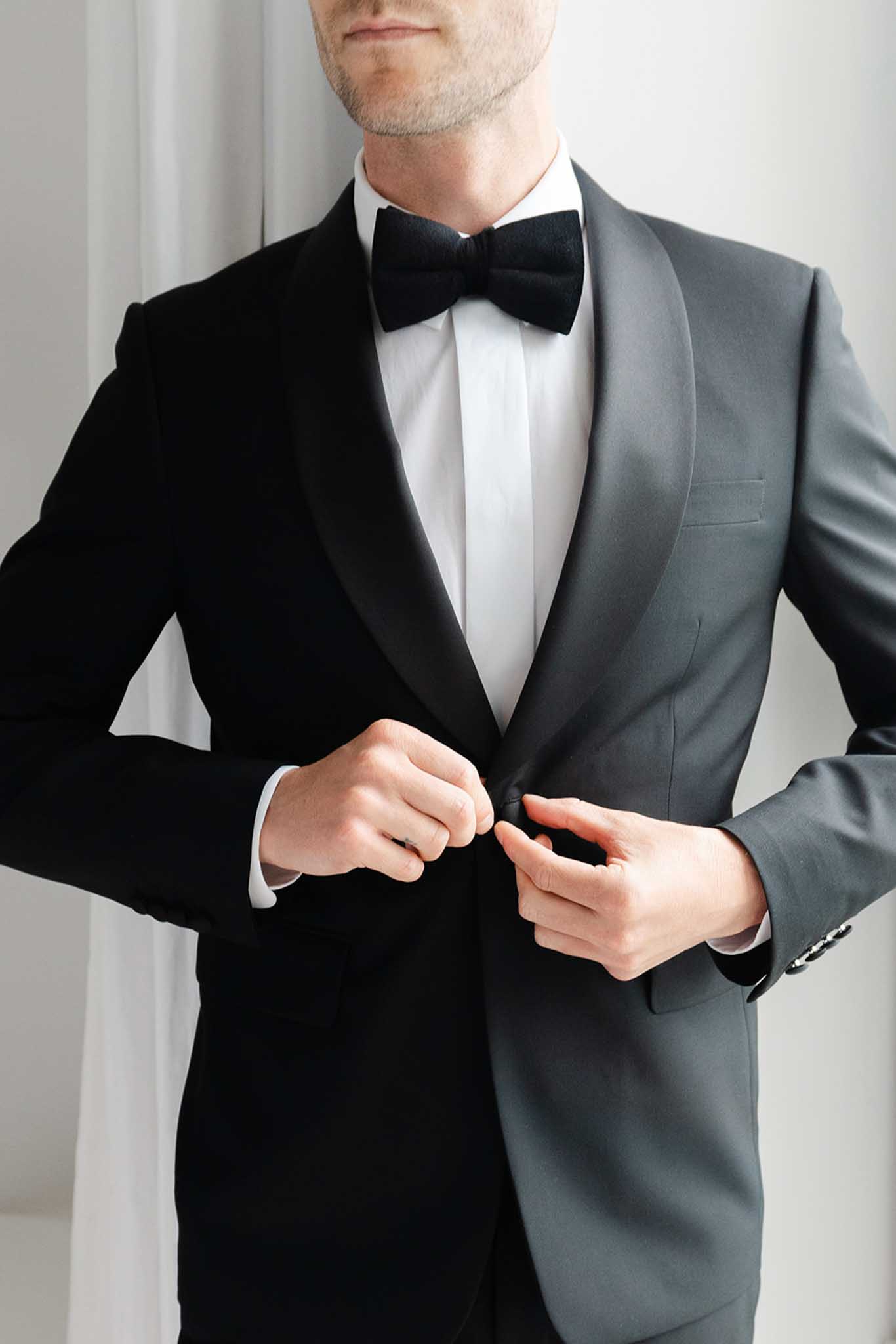 Groom adjusting black and charcoal tuxedo jacket with white shirt and black bow tie indoors