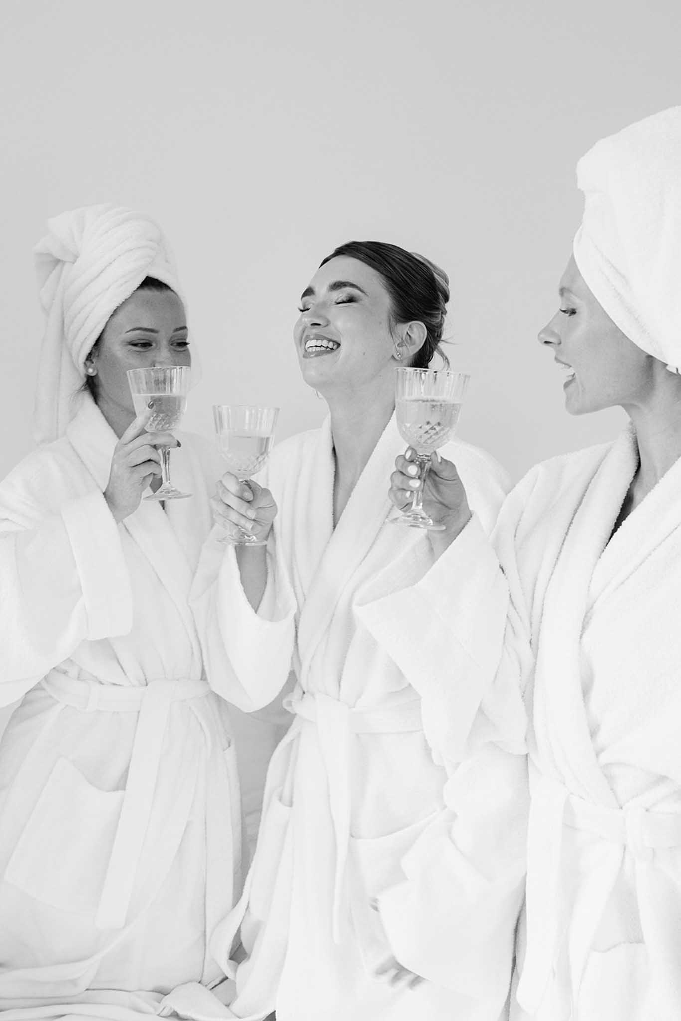 Black-and-white photo of three women in white robes laughing with champagne flutes during getting-ready moment