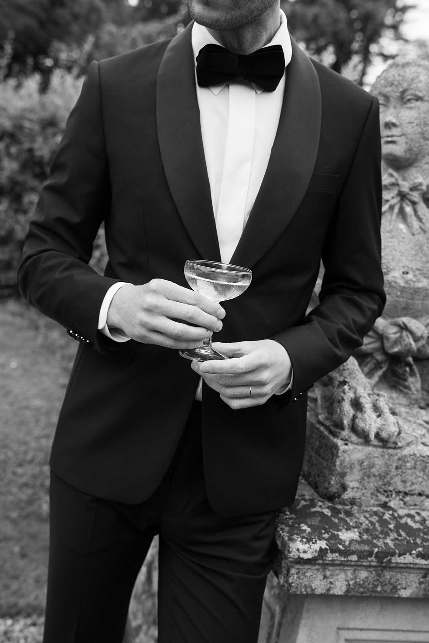 Black and white close-up of groom in tuxedo holding champagne coupe outdoors