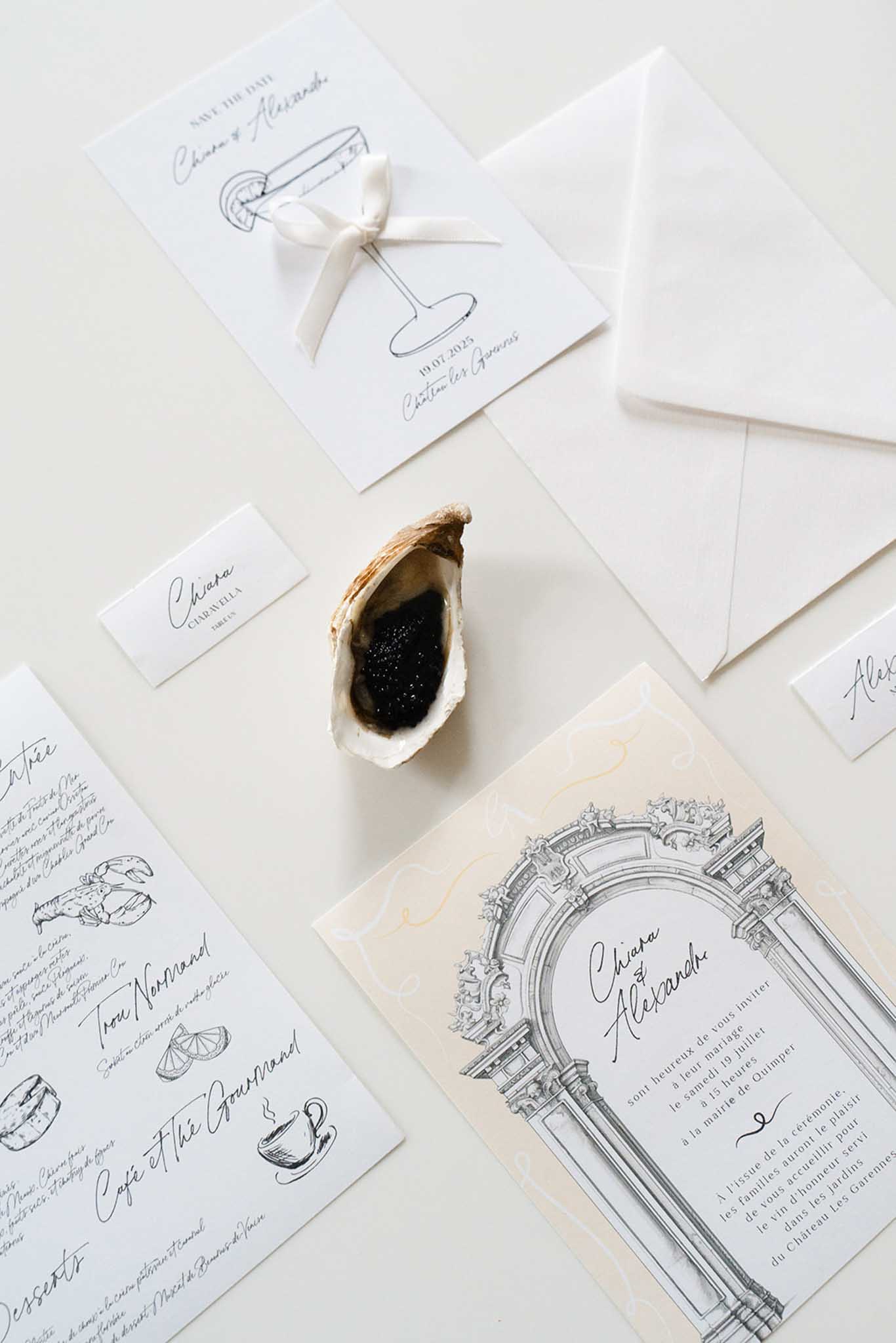 Flat lay of wedding stationery suite with black ink illustrations, cream silk ribbon, white envelopes, and oyster shell accent