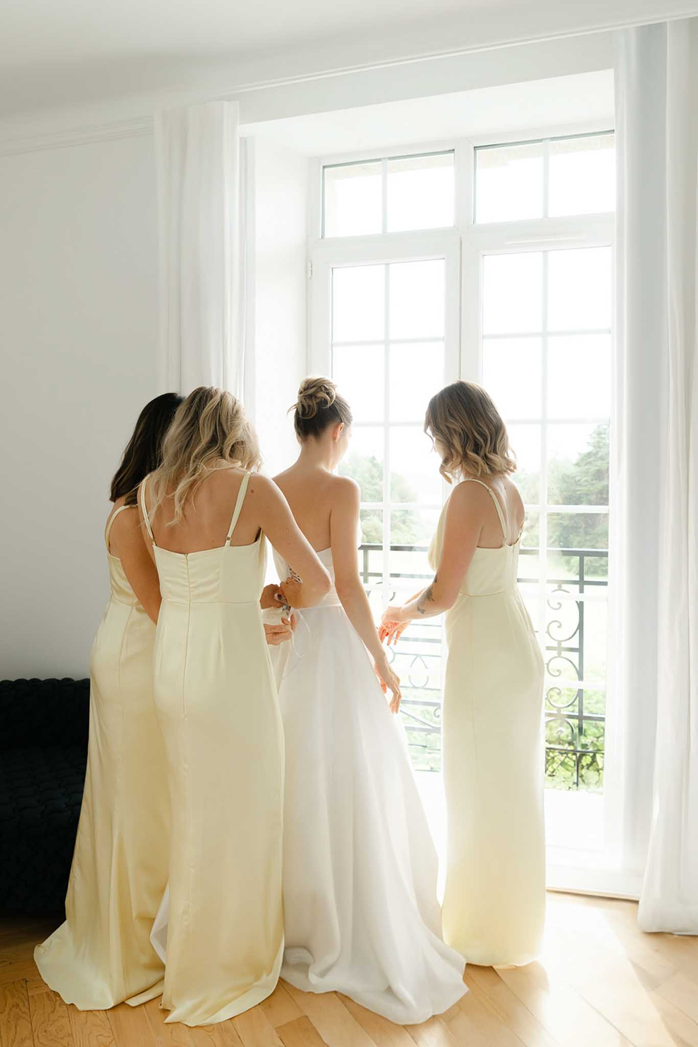 Bride in ivory gown flanked by two bridesmaids in pale yellow slip dresses at French doors, viewed from behind