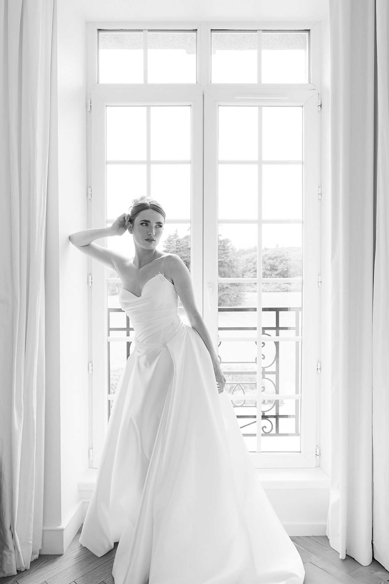 Black-and-white bridal portrait of bride in strapless gown at tall French windows with sheer curtains and ironwork beyond