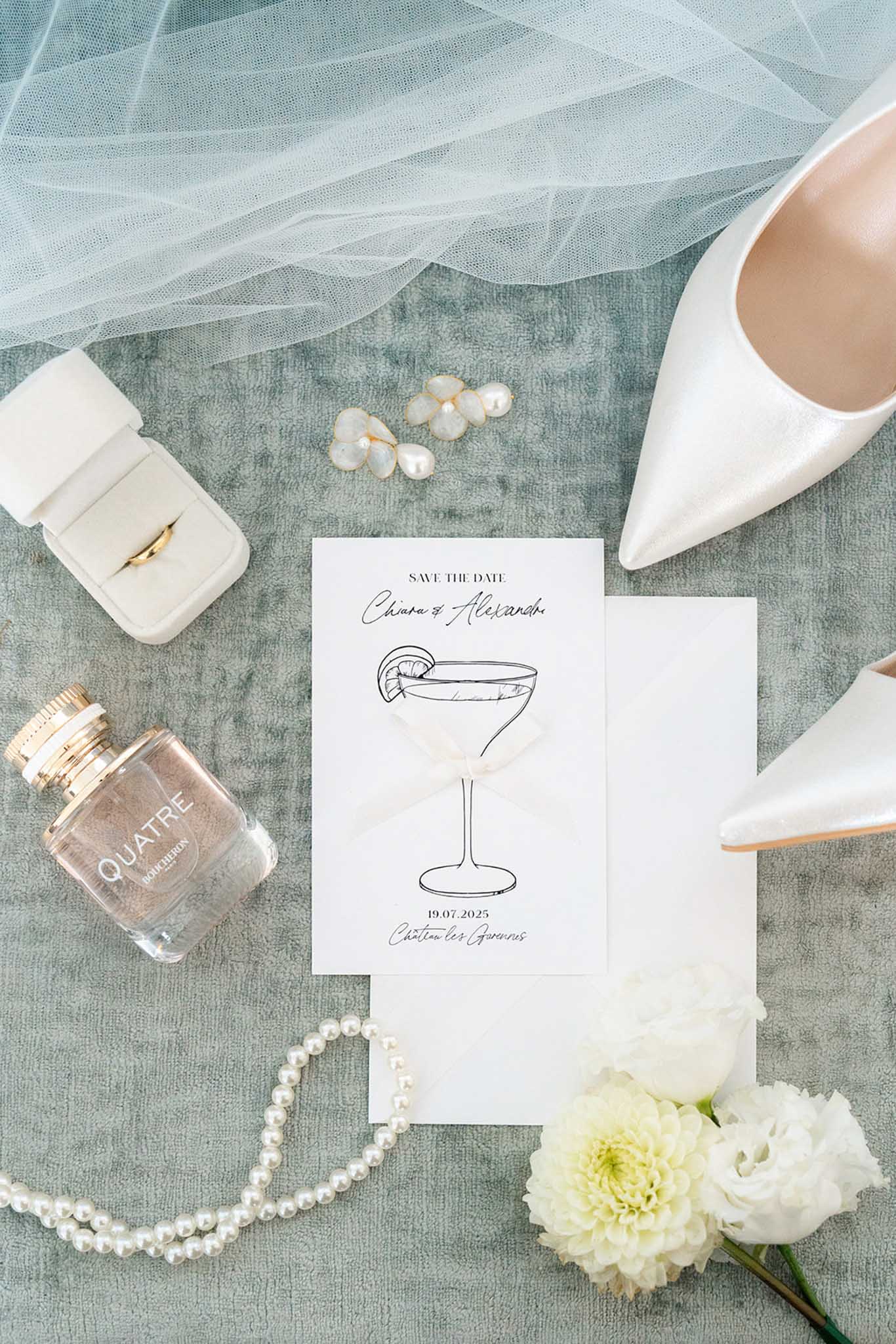 Flat lay on sage linen with save the date card, white satin shoe, pearl earrings, ring box and white dahlias