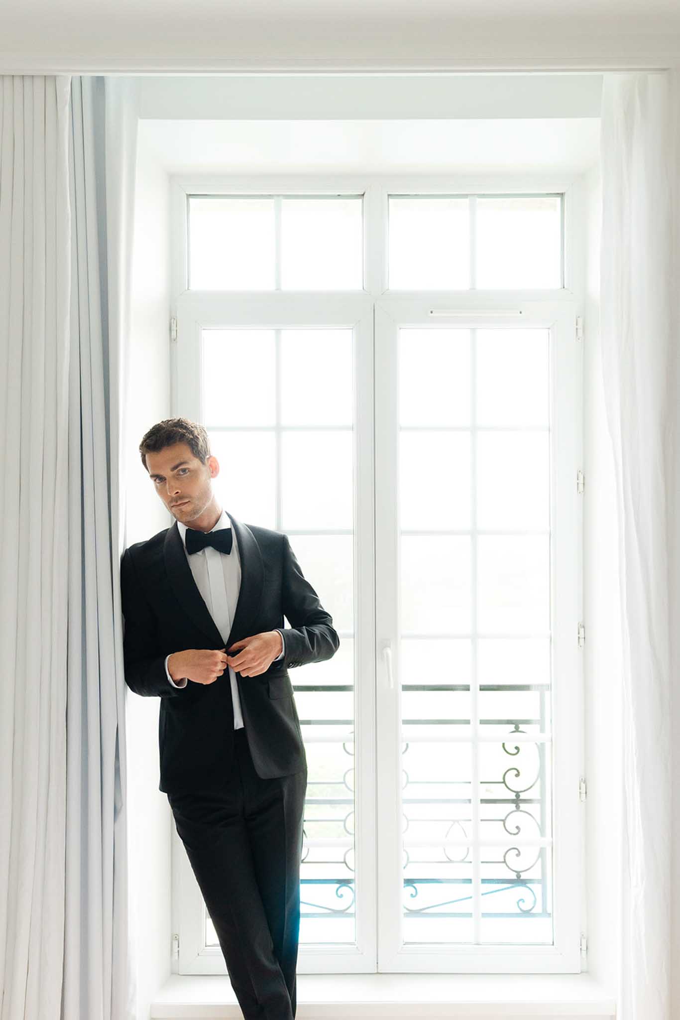 Groom fastening black tuxedo jacket in bright room with tall white French doors and balcony light