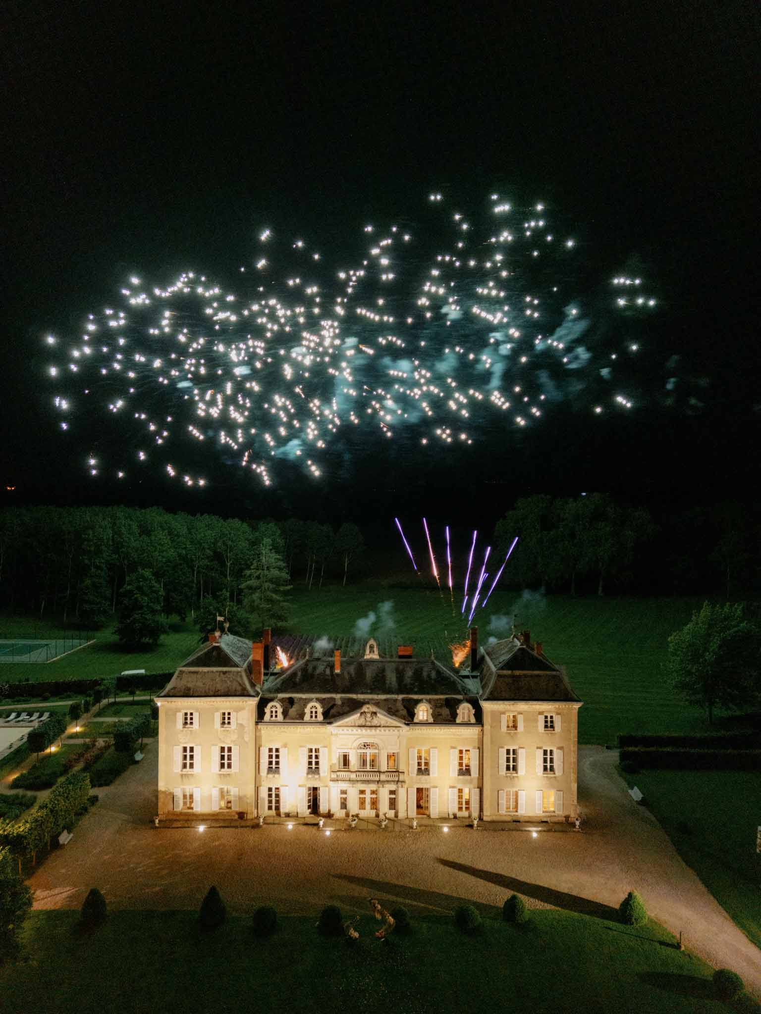Aerial night photograph of classical French château with fireworks display above illuminated mansard roof and guests in courtyard below