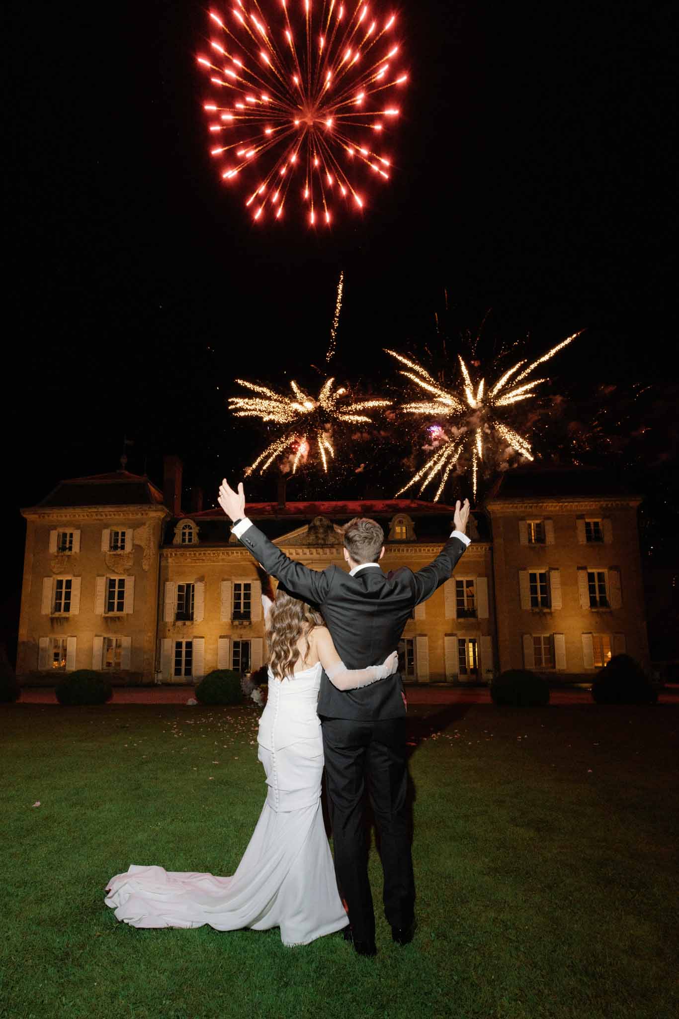 Bride and groom celebrating under red and gold fireworks at night in front of illuminated neoclassical chateau