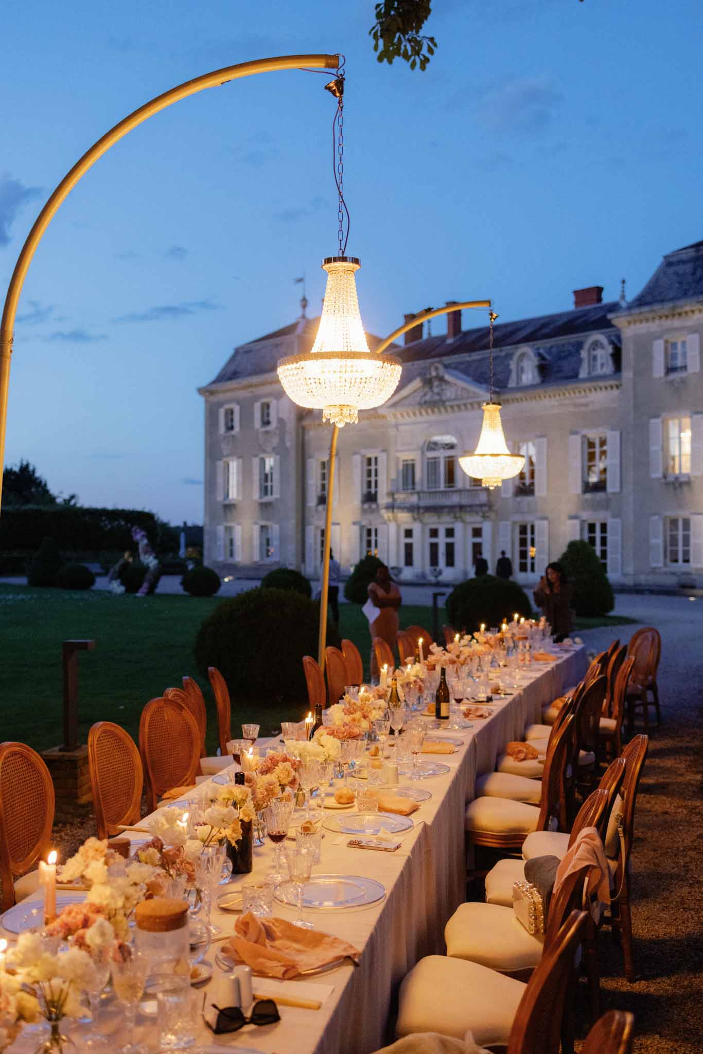 Evening banquet table on chateau grounds with rattan chairs, crystal chandeliers on gold stands at dusk