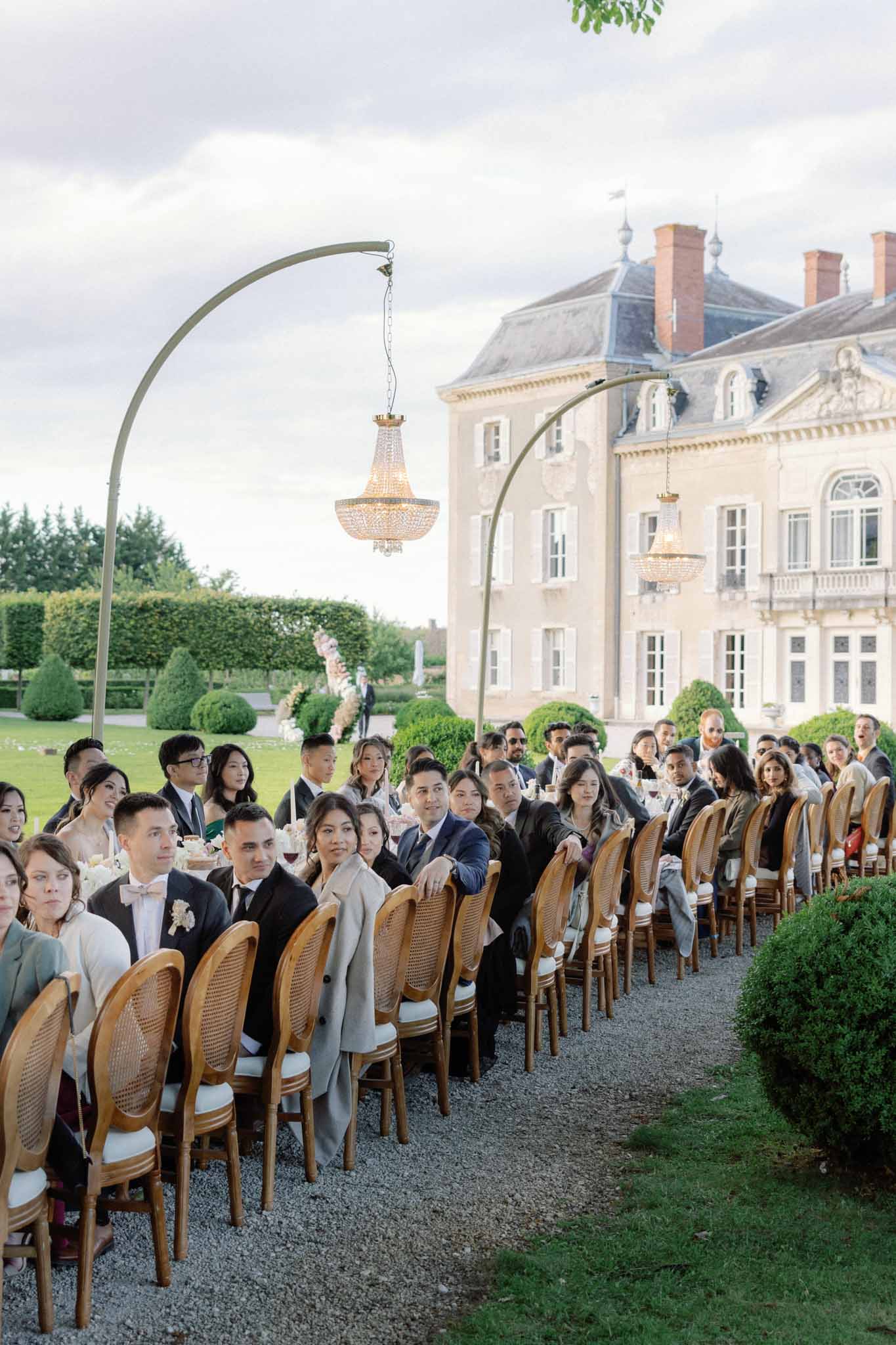 Outdoor ceremony on gravel path at French chateau with seated guests, golden chandeliers on arches, and topiary gardens