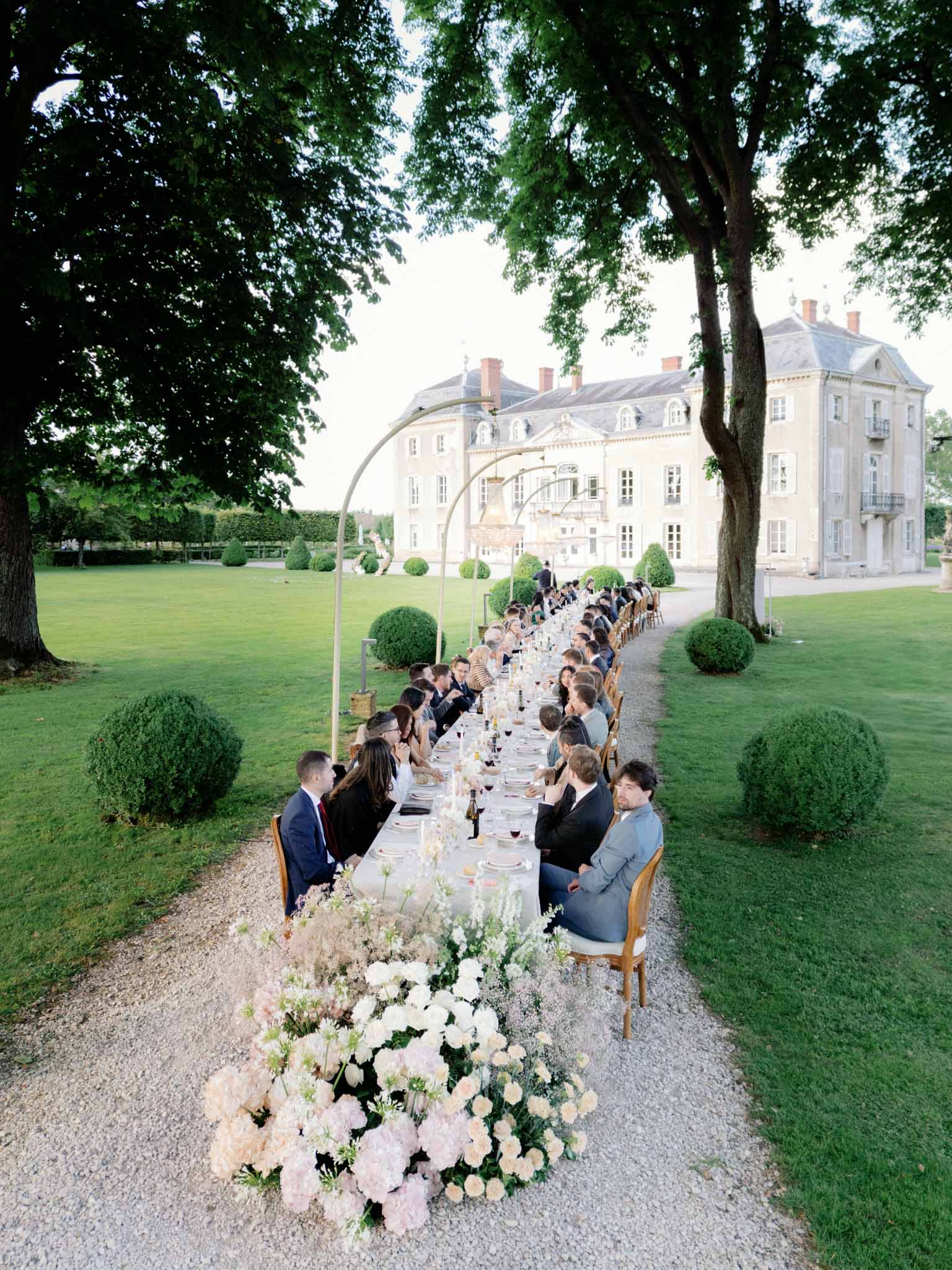 Aerial view of outdoor reception with 40-50 guests at extended table on chateau grounds, cream stone manor at far end