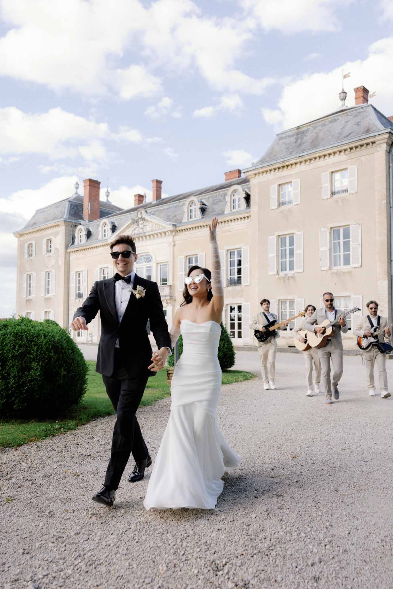 Bride and groom walking down gravel driveway in front of cream chateau with mansard roofs