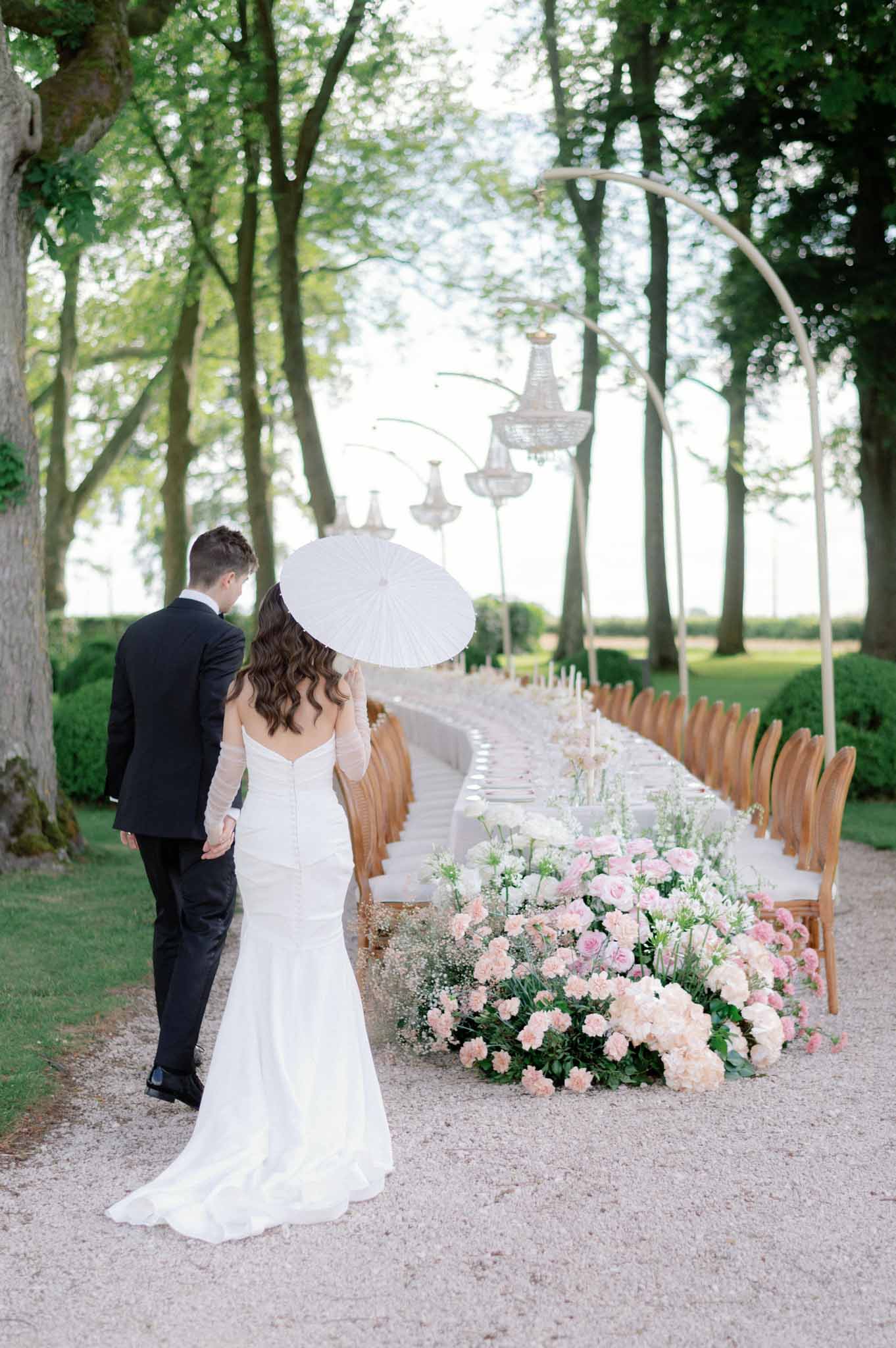 Bride with white parasol and groom walking down a tree-lined aisle decorated with pink carnations