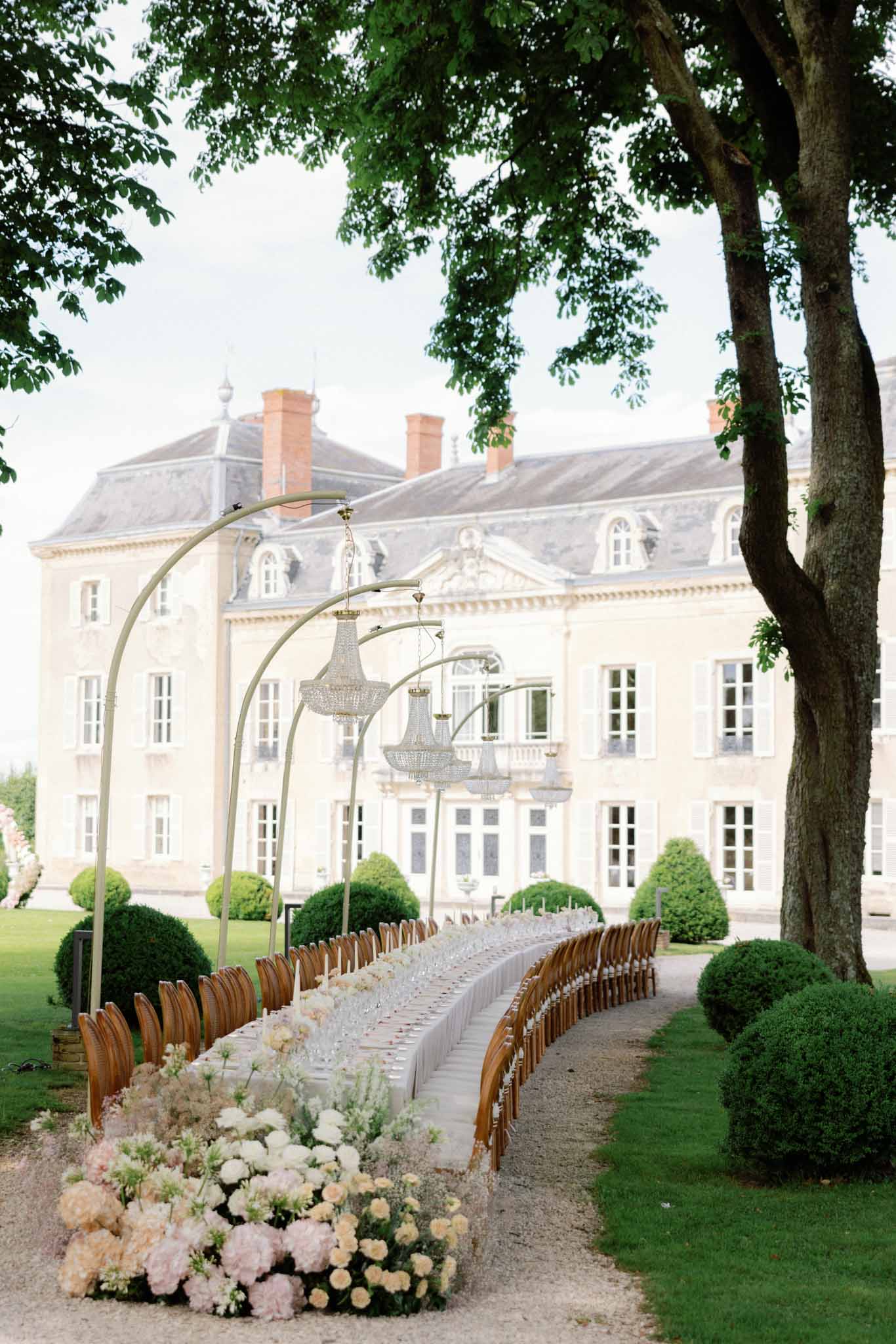 Ceremony aisle lined with wood chairs, pale pink hydrangeas and crystal chandelier arbors, classical château visible behind.