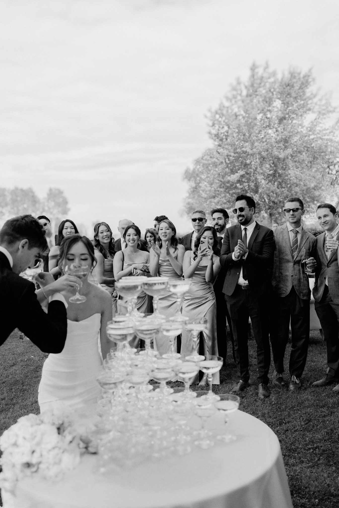 Black and white photo of bride and groom raising champagne glasses during outdoor toasts with guests gathered around