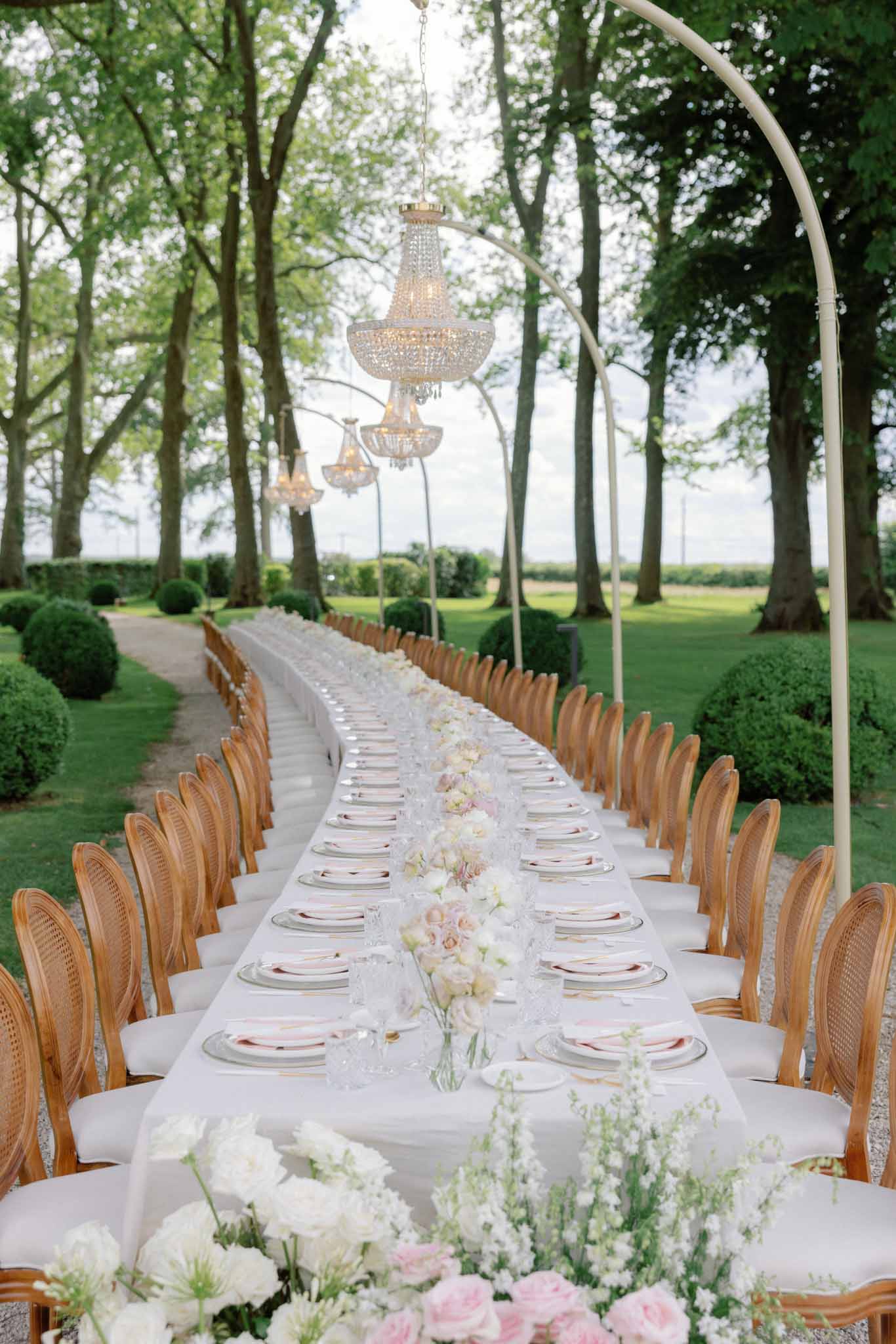 Outdoor reception table in tree-lined garden alley with white and pink floral runner and crystal chandelier pendants