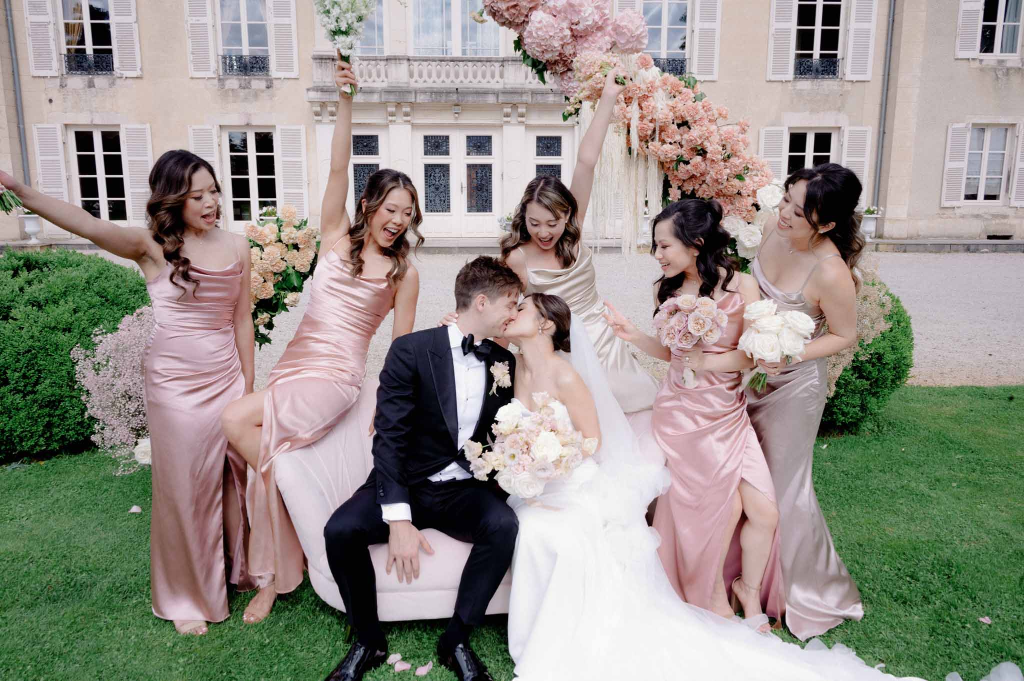 Bride, groom and bridesmaids in dusty rose gowns posing in château courtyard under cascading floral installations