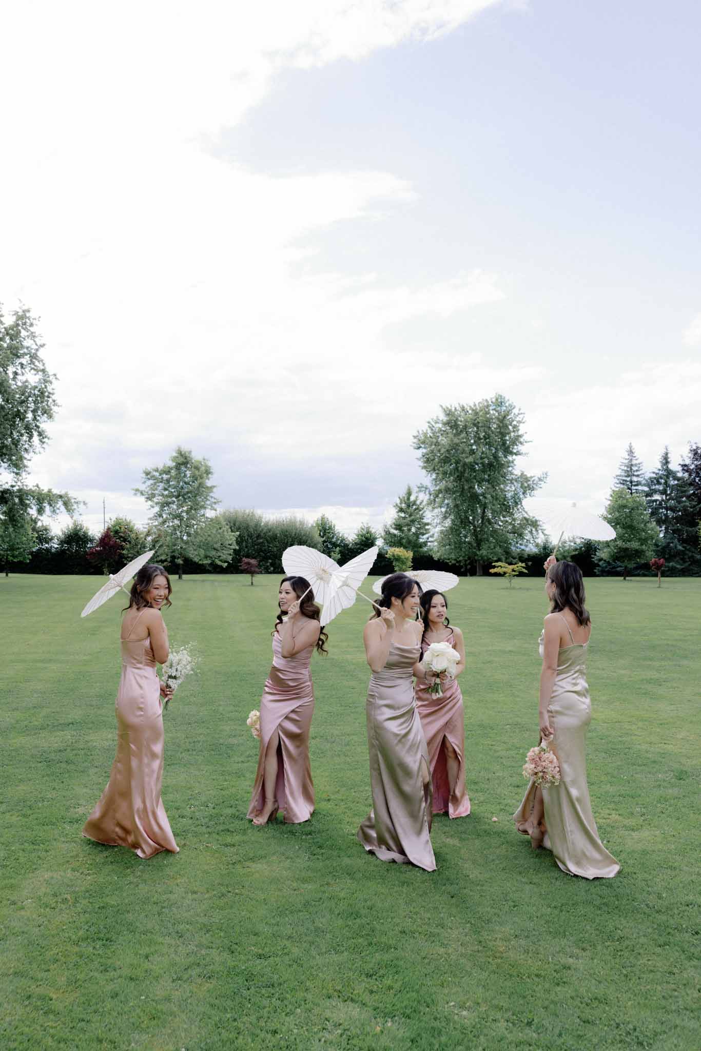 Five bridesmaids in dusty rose and taupe gowns on lawn, three with white umbrellas and angel wings, two with bouquets
