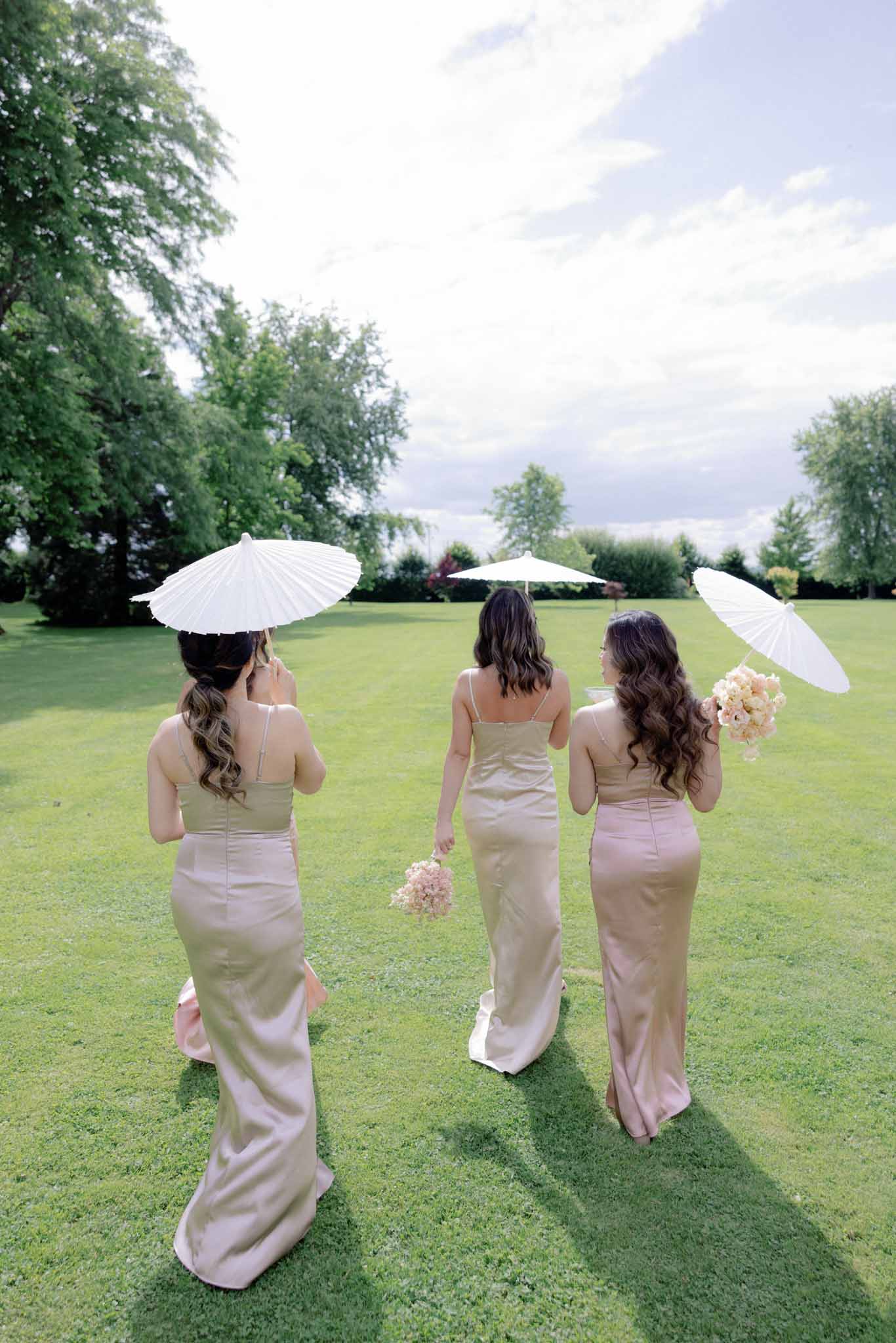 Three bridesmaids in champagne and dusty rose gowns walking across an estate lawn, each holding a white parasol.