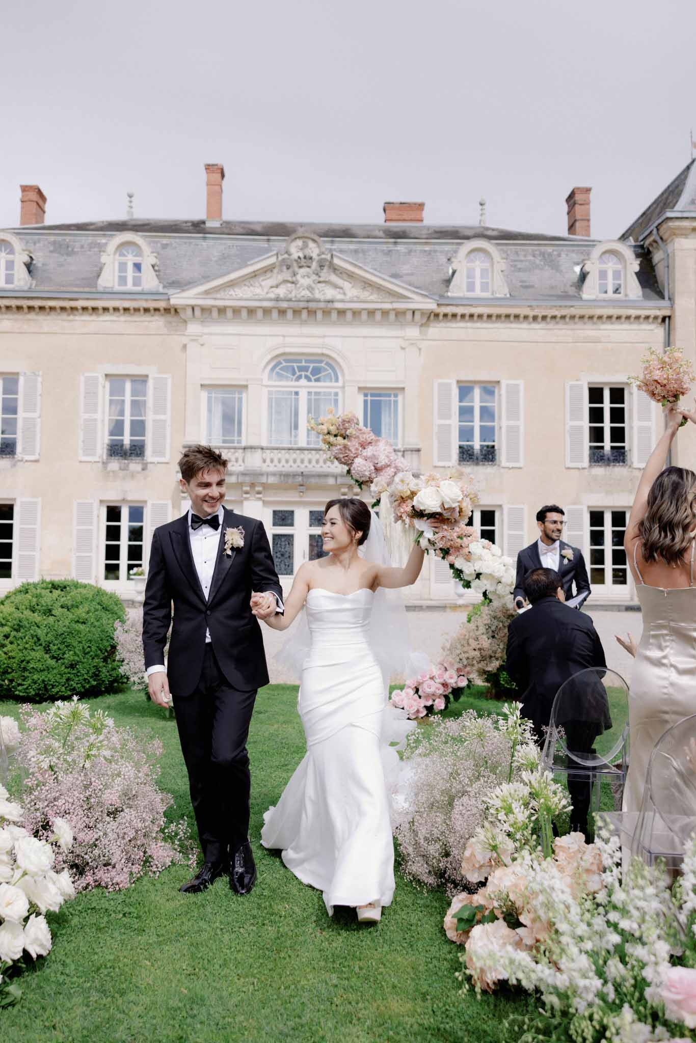 Bride and groom walking down aisle in chateau courtyard, white and dusty pink floral installations flanking, manor behind
