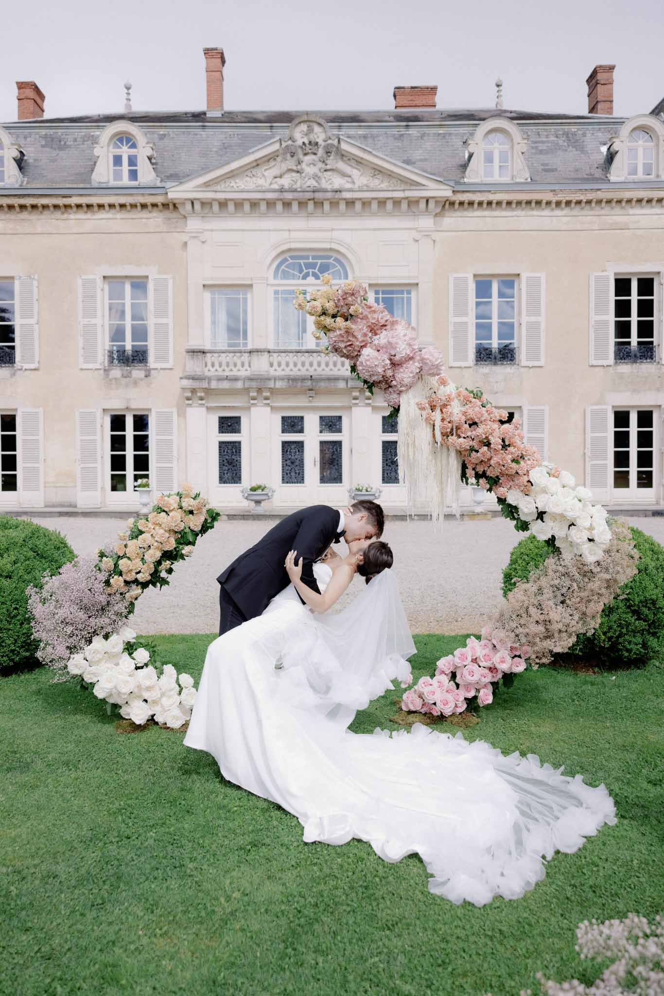 Groom dipping bride on chateau lawn framed by dusty rose and blush floral installations