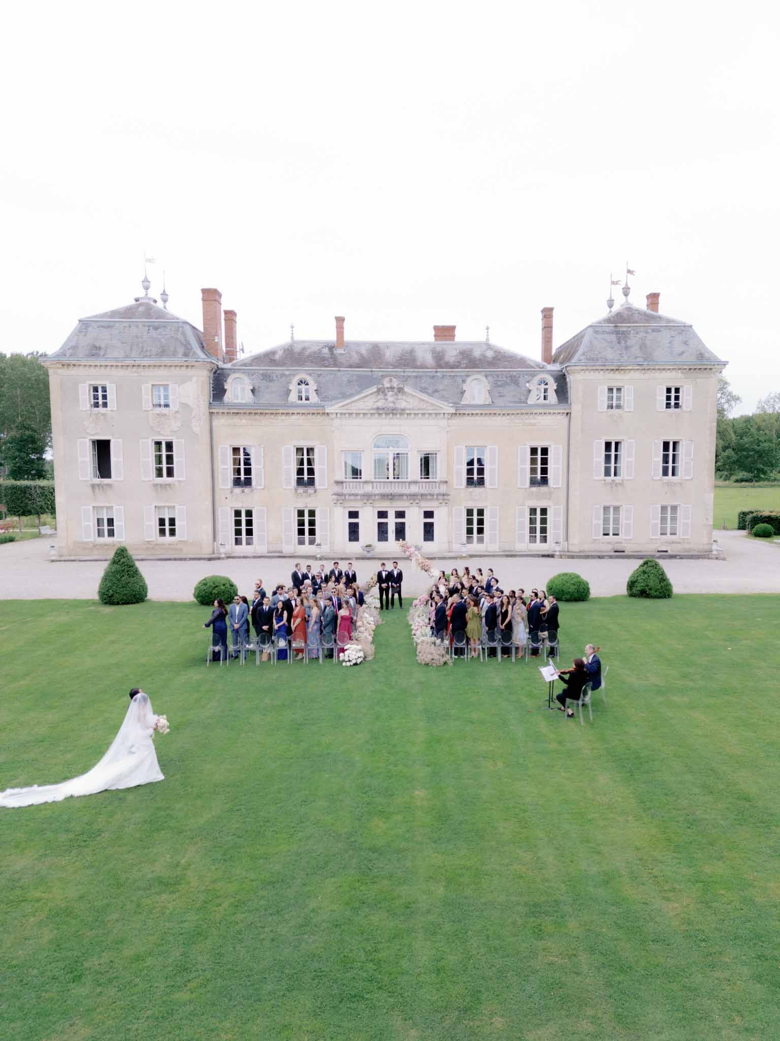 Aerial view of wedding ceremony on chateau lawn with guests in semi-circle around white floral altar