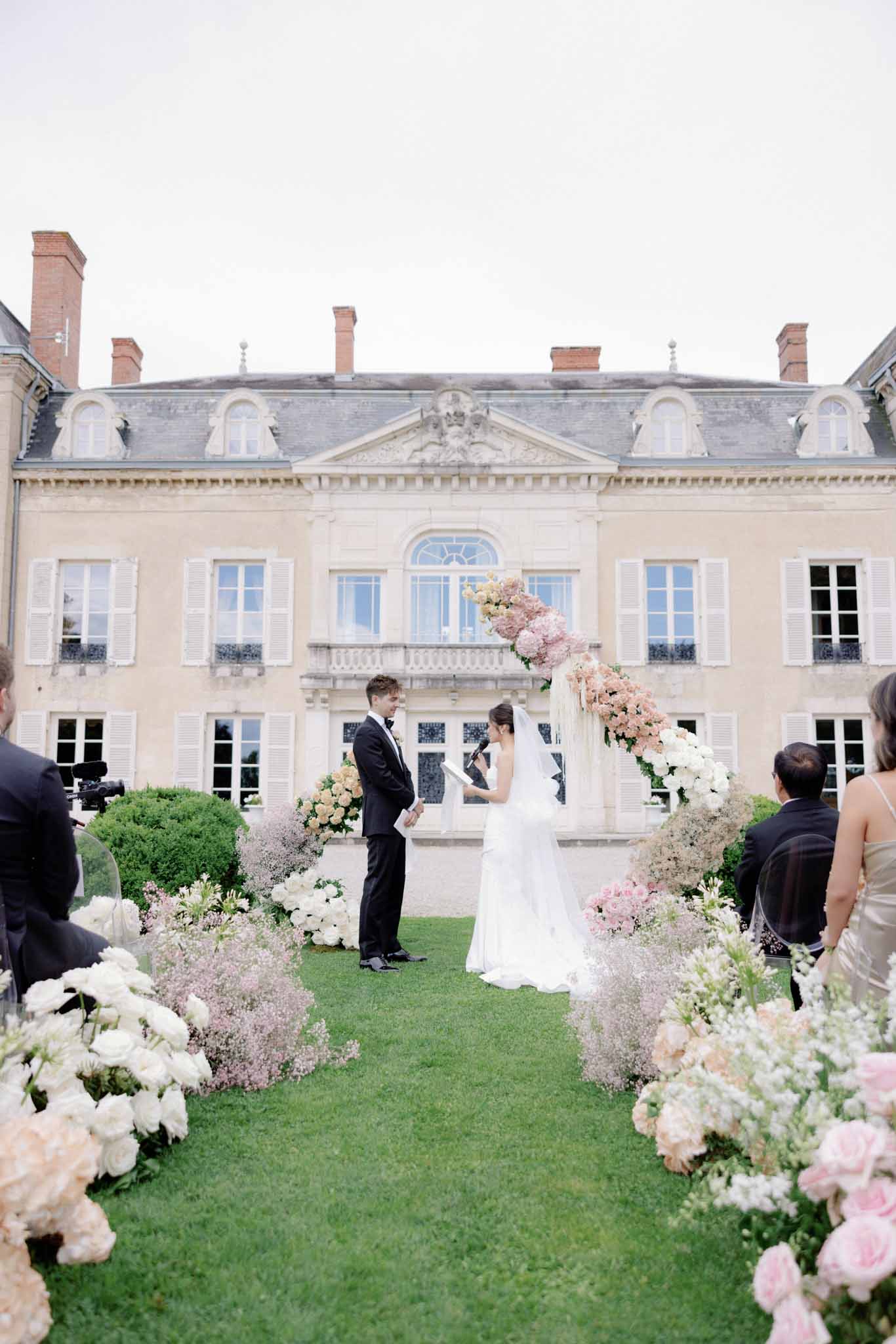 Wedding ceremony on château lawn with couple beneath blush and white floral arch, guests lining aisle of rose and hydrangea clusters