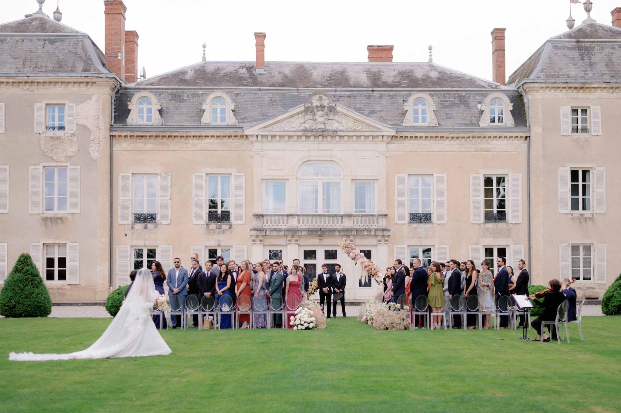 Outdoor ceremony on chateau lawn with guests in grey chairs and blush floral arrangements