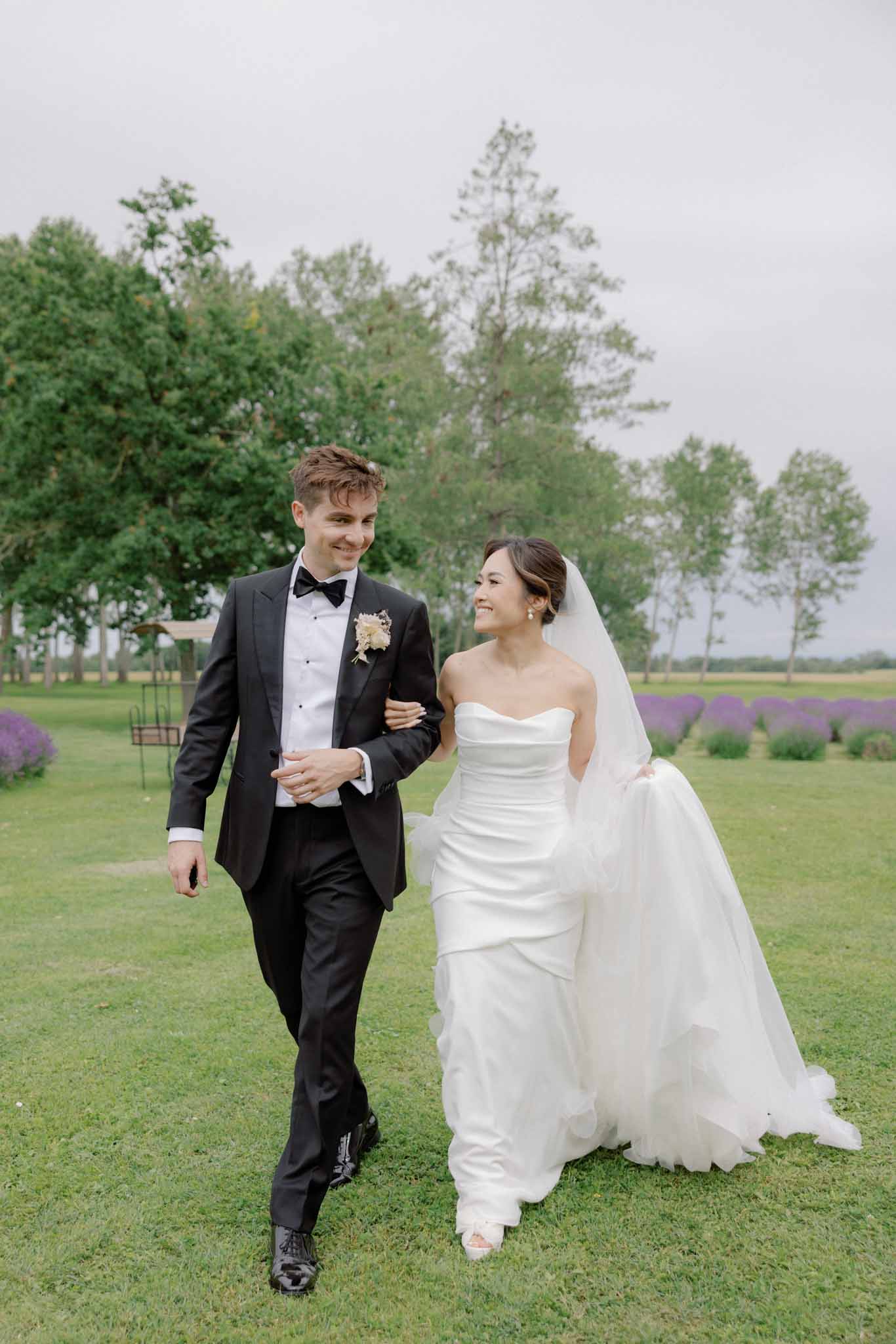Bride in ivory strapless gown with tulle veil and groom in black tuxedo walk across lawn with lavender rows behind