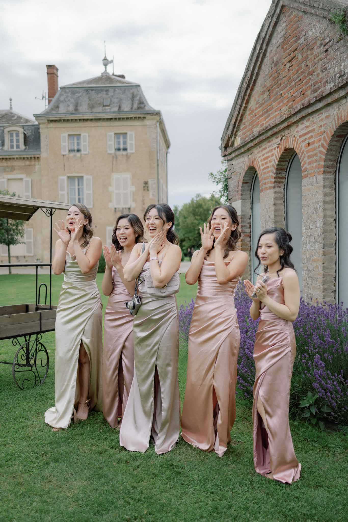 Five bridesmaids in blush and champagne satin gowns laughing on lawn in front of brick chateau with lavender plants