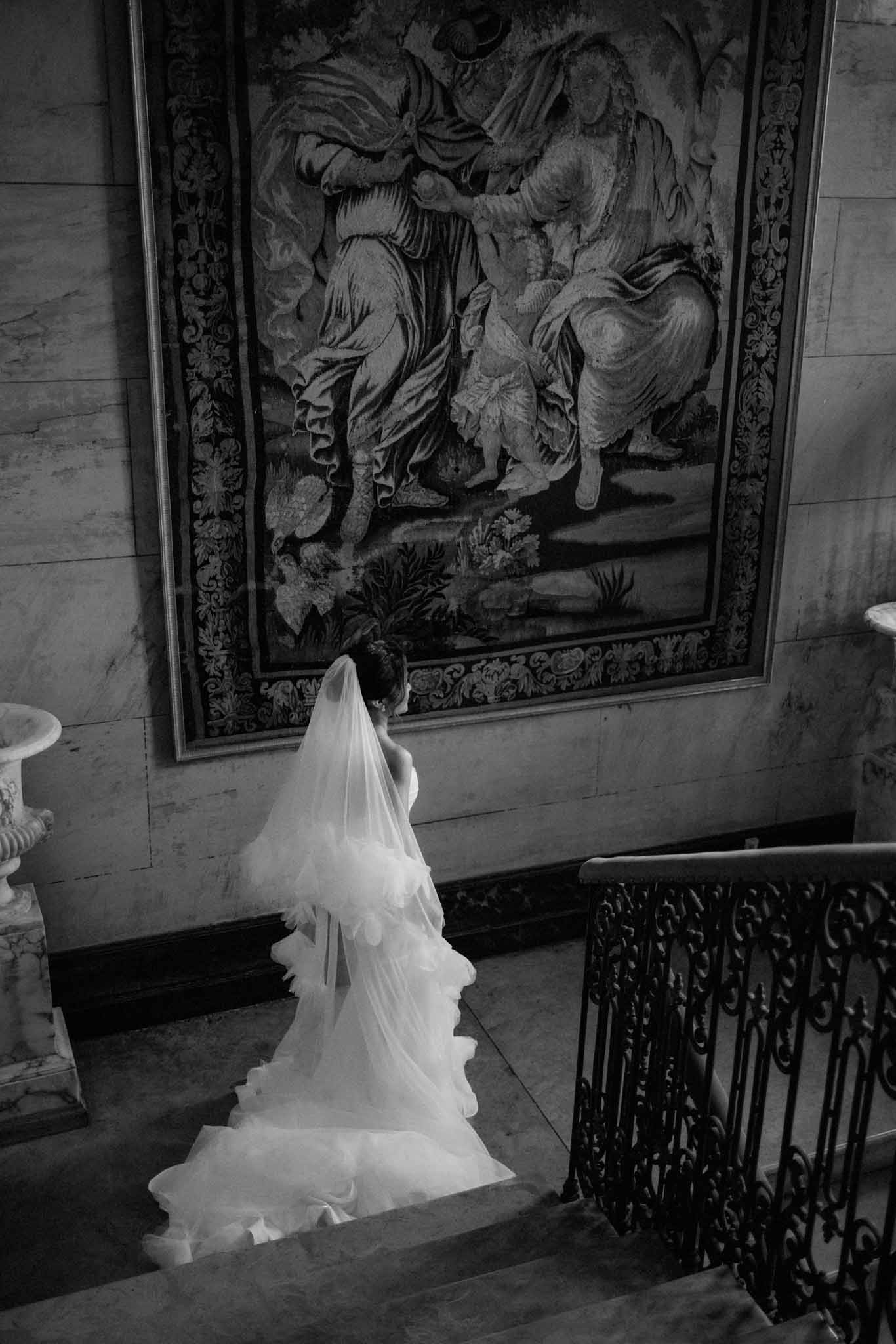 Bride from behind in ivory gown and long veil, looking up at classical painting in ornate neoclassical hall