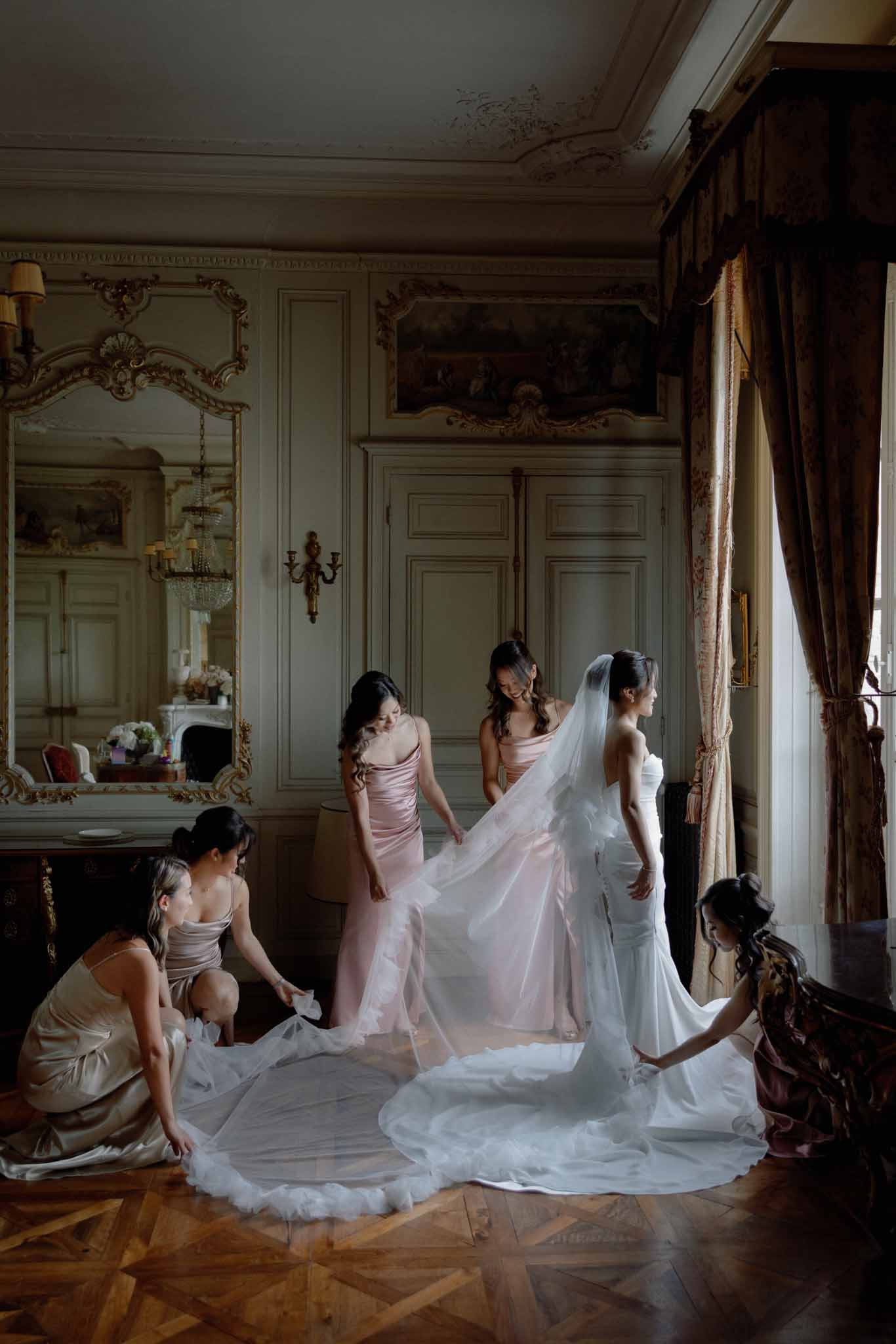 Bride surrounded by six bridesmaids in dusty rose dresses arranging her veil in a neoclassical salon