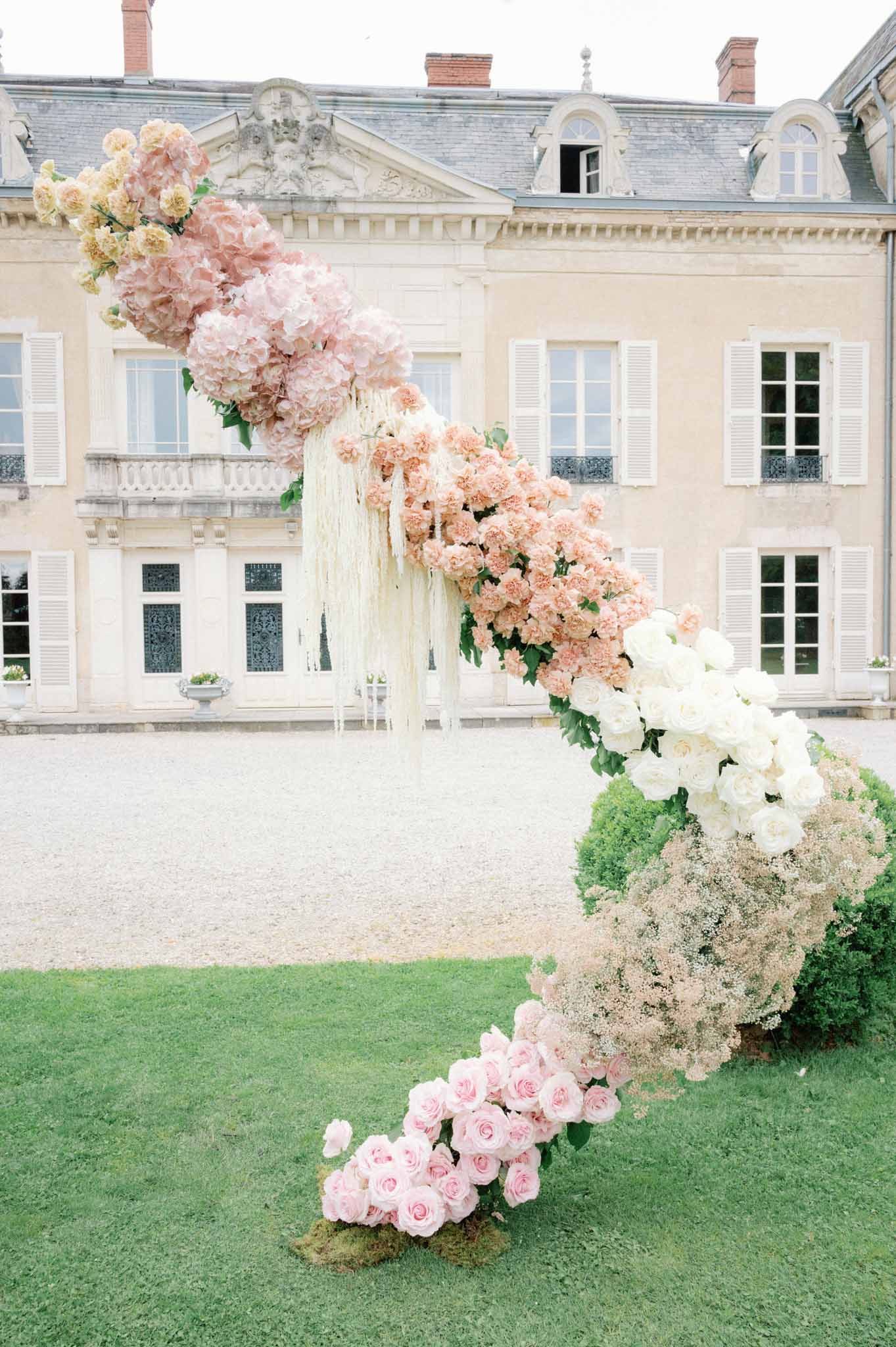 Cascading floral installation in pink and ivory on lawn before classical French chateau facade