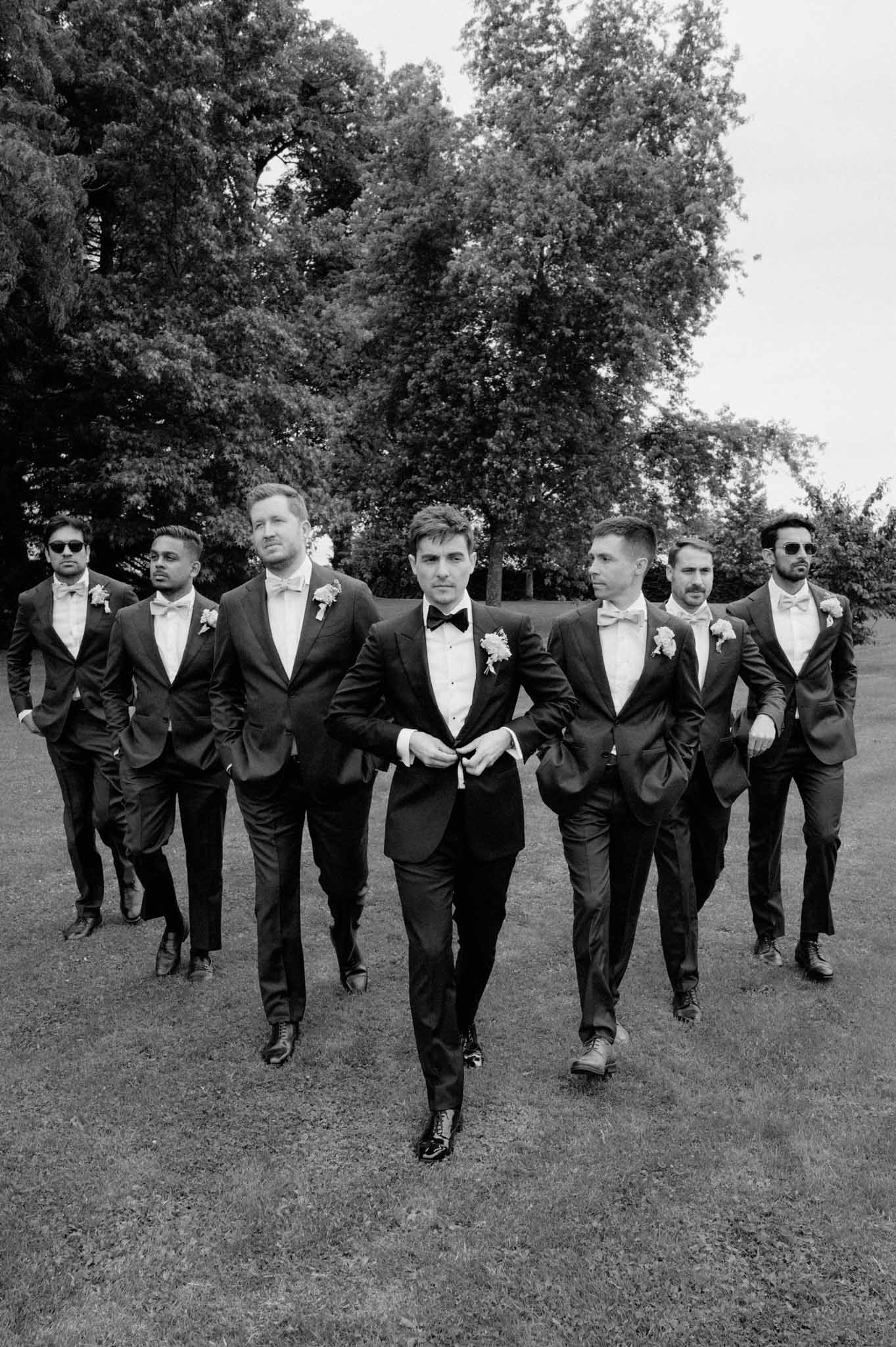Groom and seven groomsmen in black suits walk tree-lined garden avenue with church spire visible, black-and-white