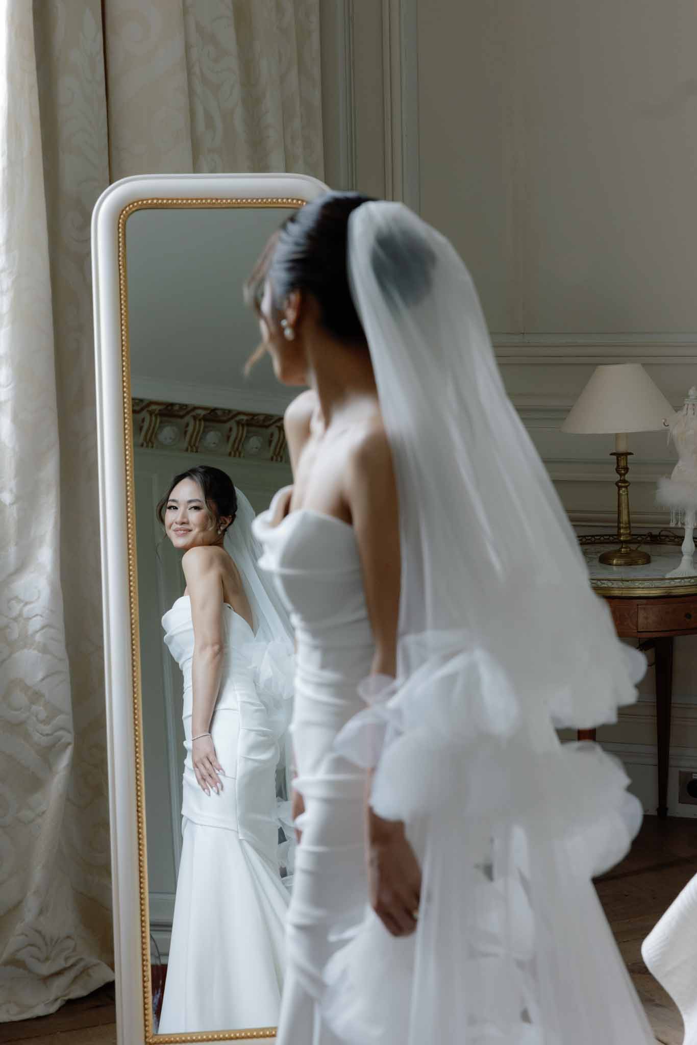Bride viewing her reflection in an ornate gold-framed mirror in a classical room with sage green panelled walls; white strapless gown.