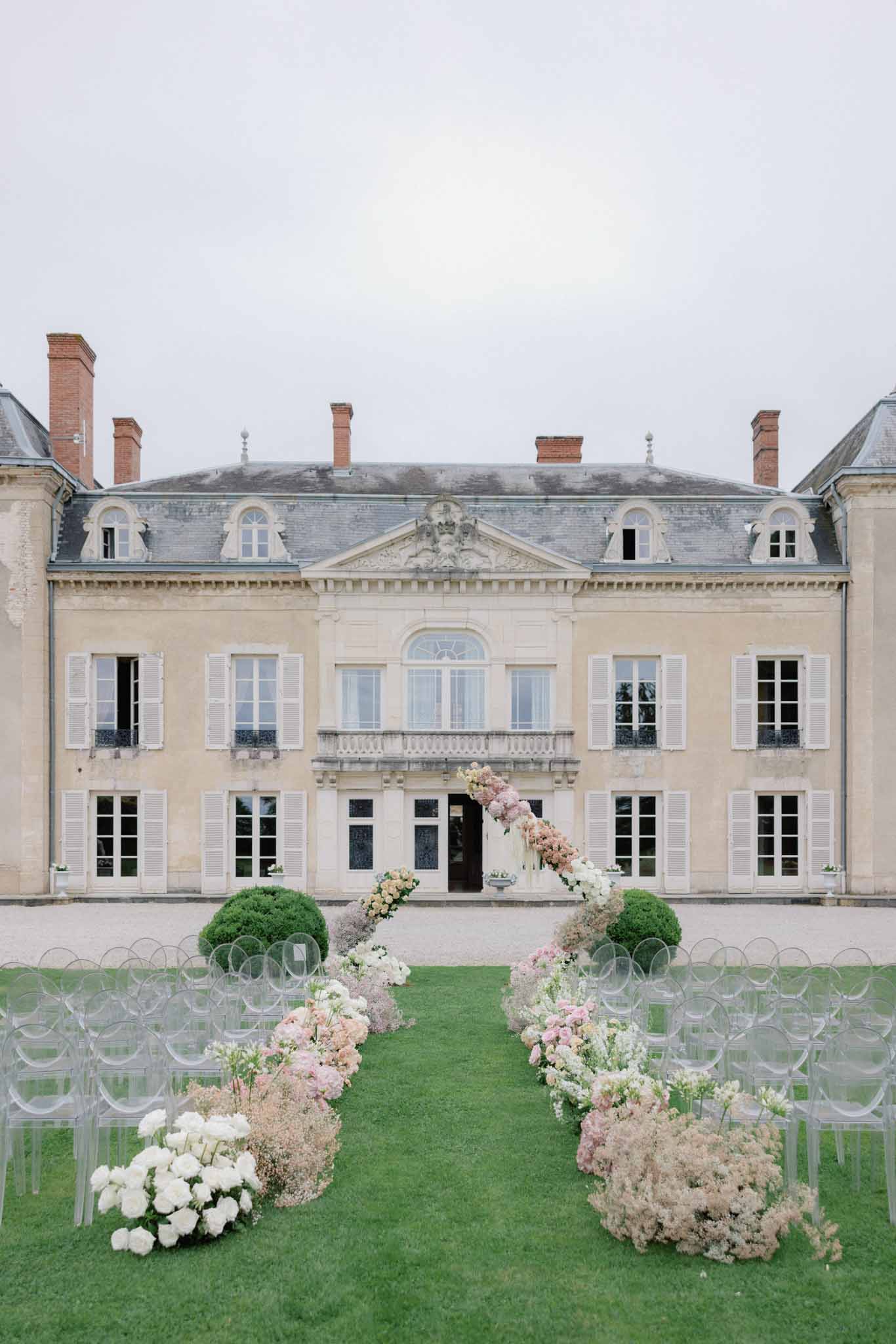 Ceremony aisle lined with blush roses and white hydrangeas in transparent acrylic chairs in courtyard of French château