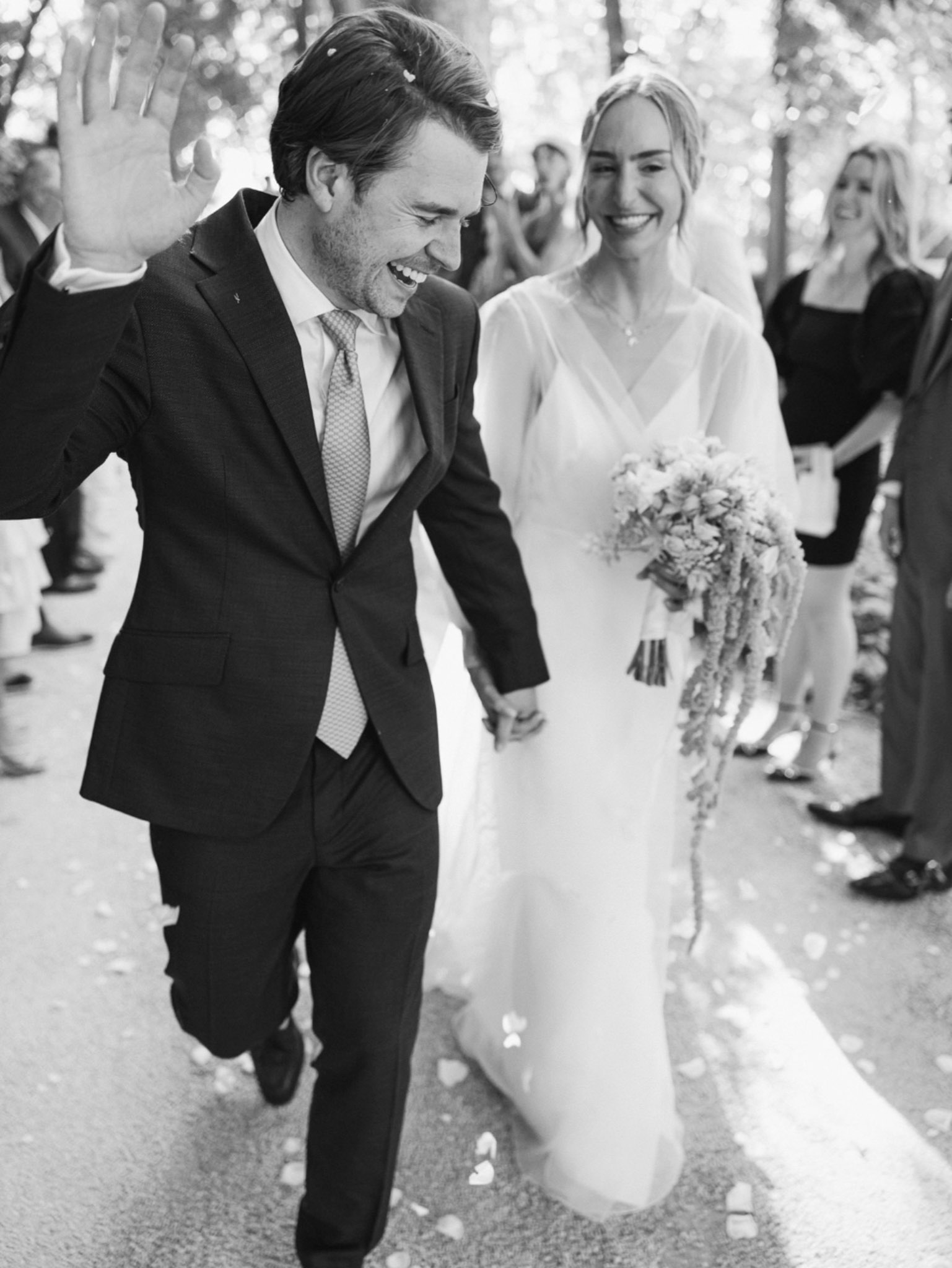 Bride and groom walking hand-in-hand down the aisle as guests wave and celebrate in black and white