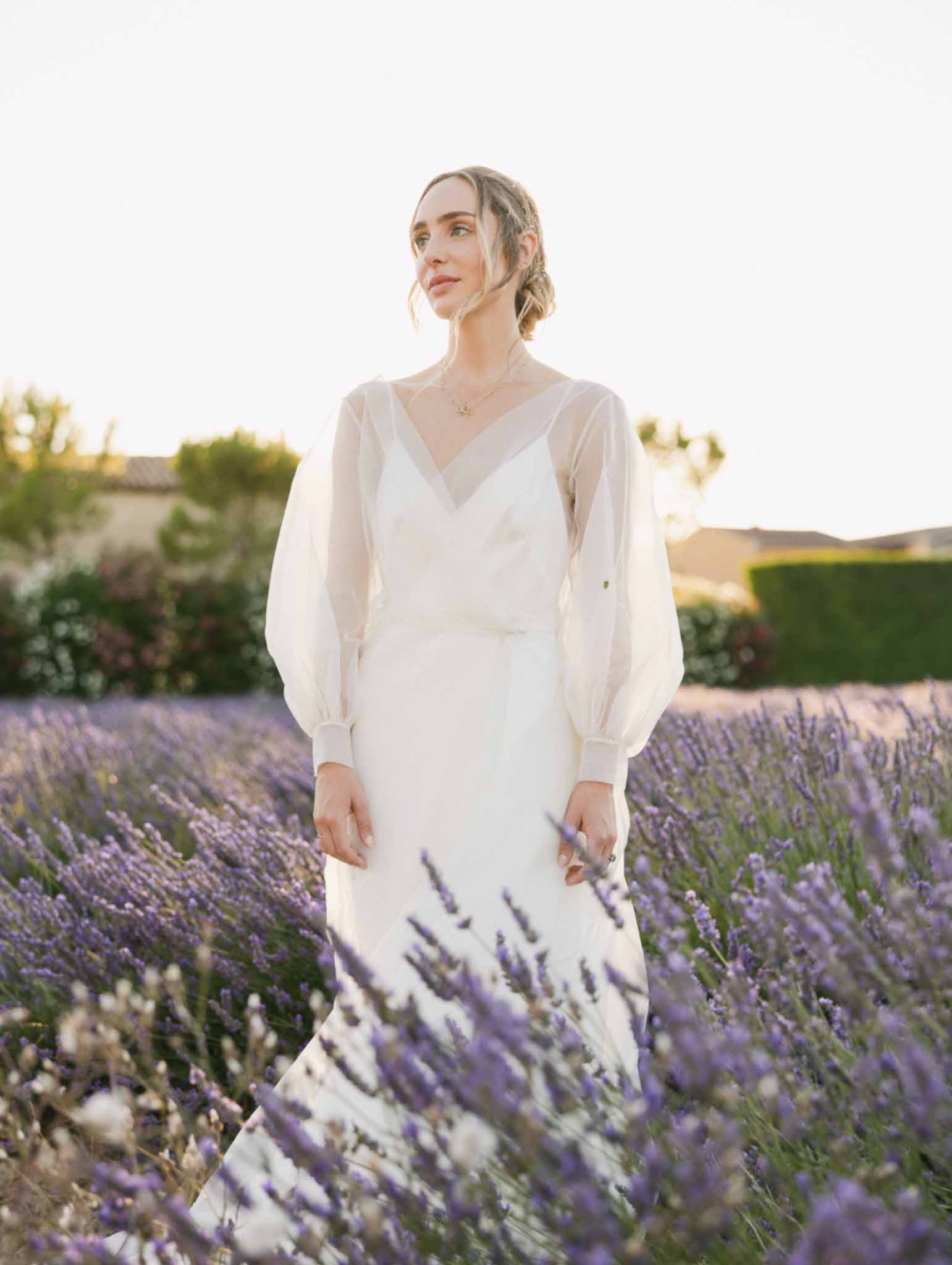 Bride in ivory silk wrap dress stands in a lavender field with Provencal stone buildings in the background