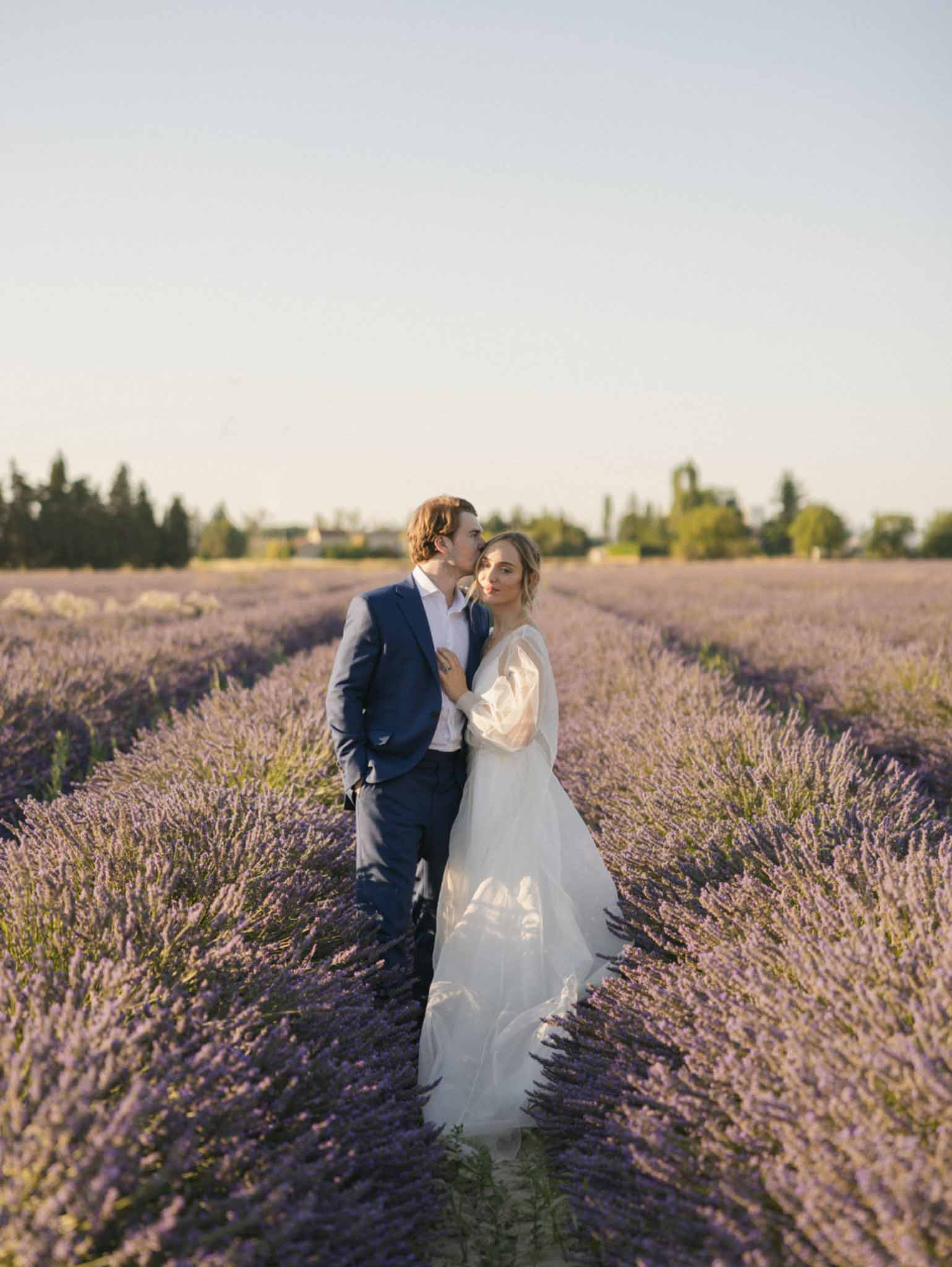 Couple posing in lavender field, bride in long-sleeved ivory gown and groom in navy suit