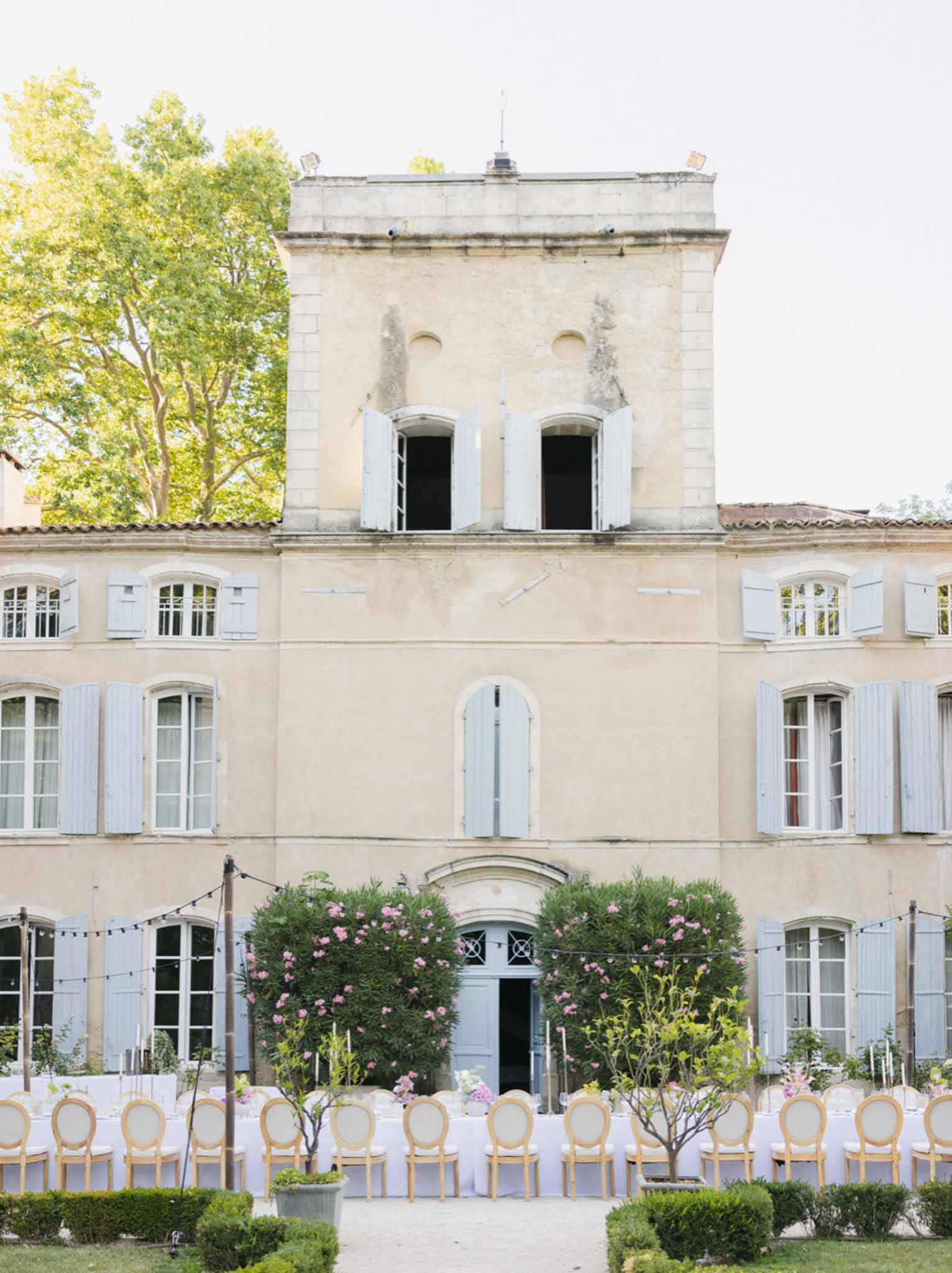 Ceremony courtyard at cream French stone mansion with pink rose topiaries, round-back chairs, and string lighting