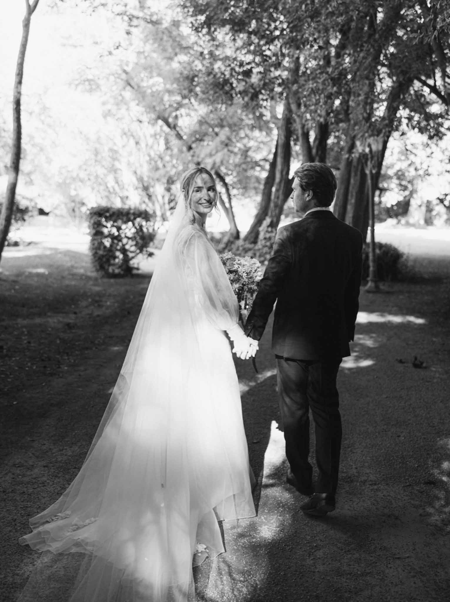 Bride in white gown with veil looks toward camera holding hands with groom on tree-lined avenue, black-and-white