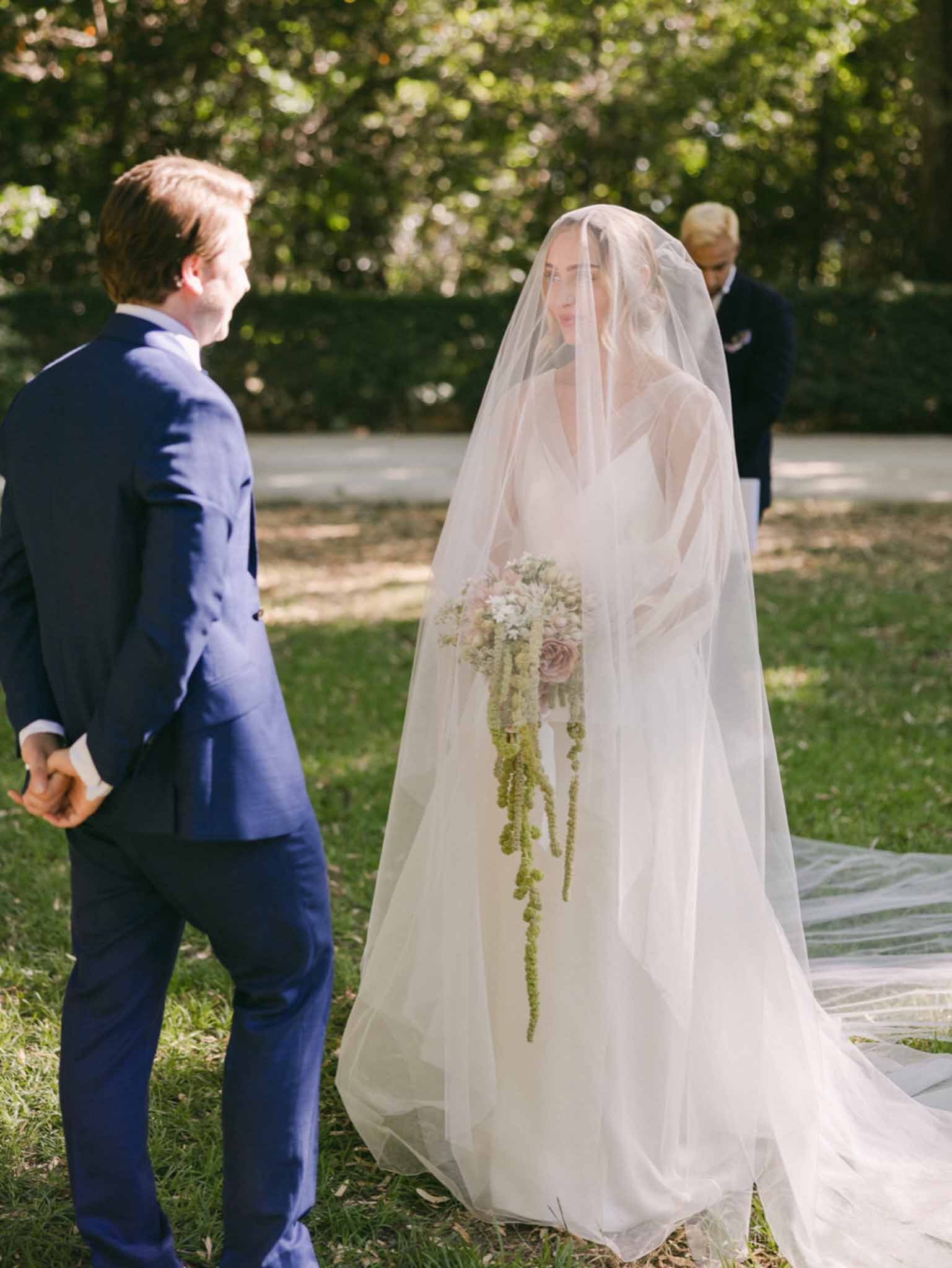 Bride with trailing bouquet and full veil facing groom in navy suit during outdoor garden ceremony