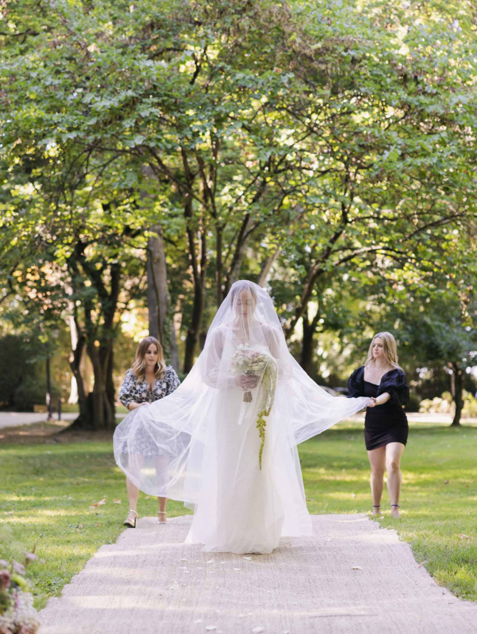 Bride walking down tree-lined garden path with trailing veil and cascading bouquet accompanied by two attendants