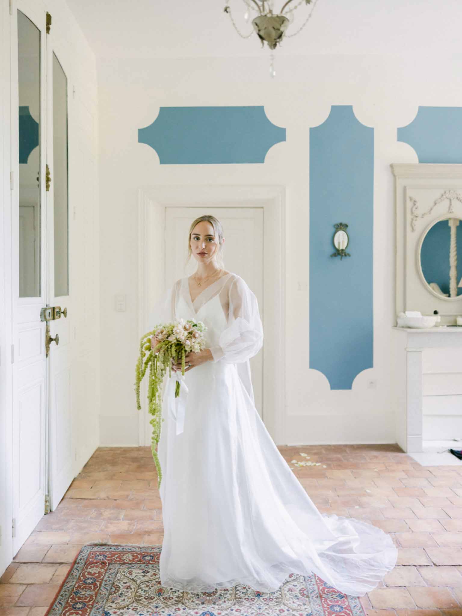 Bride in ivory long-sleeve V-neck gown holding cascading bouquet in hallway with terracotta tile floors and blue geometric wall panels