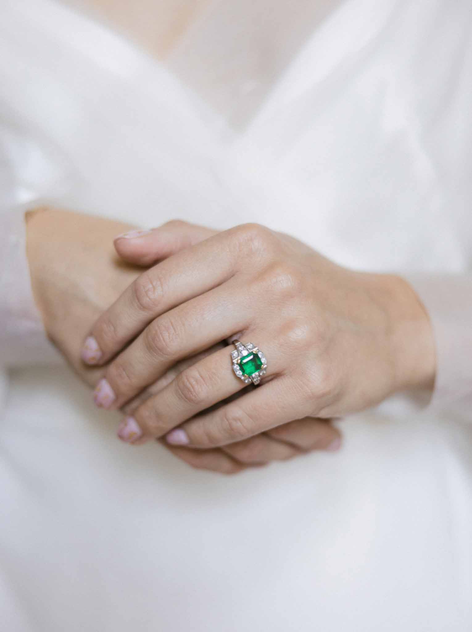 Close-up of newlyweds' hands showing emerald-cut green gemstone engagement ring with diamond accents and plain white metal wedding band