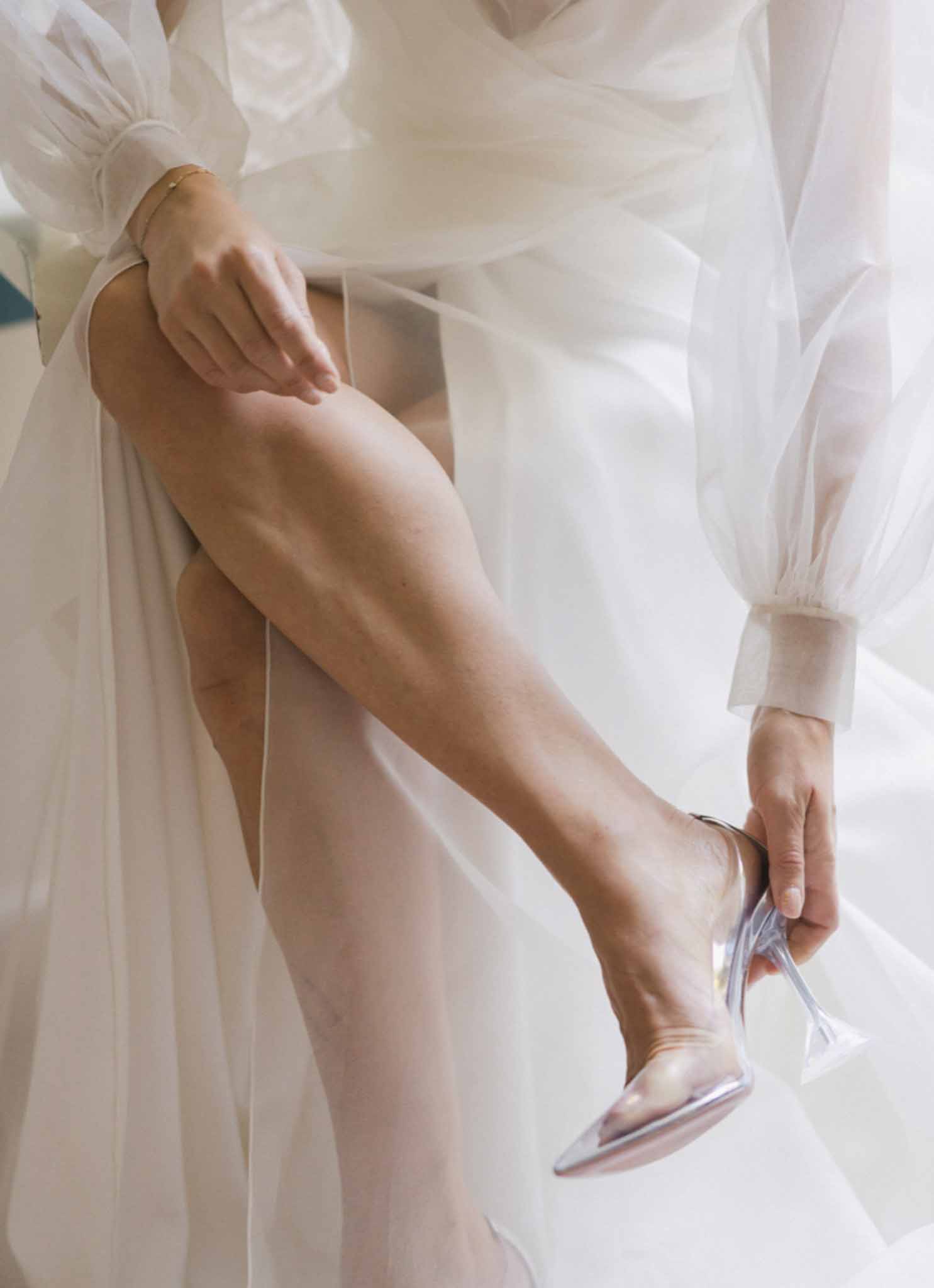 Close-up of bride adjusting silver flat shoes with flowing ivory dress fabric during getting ready